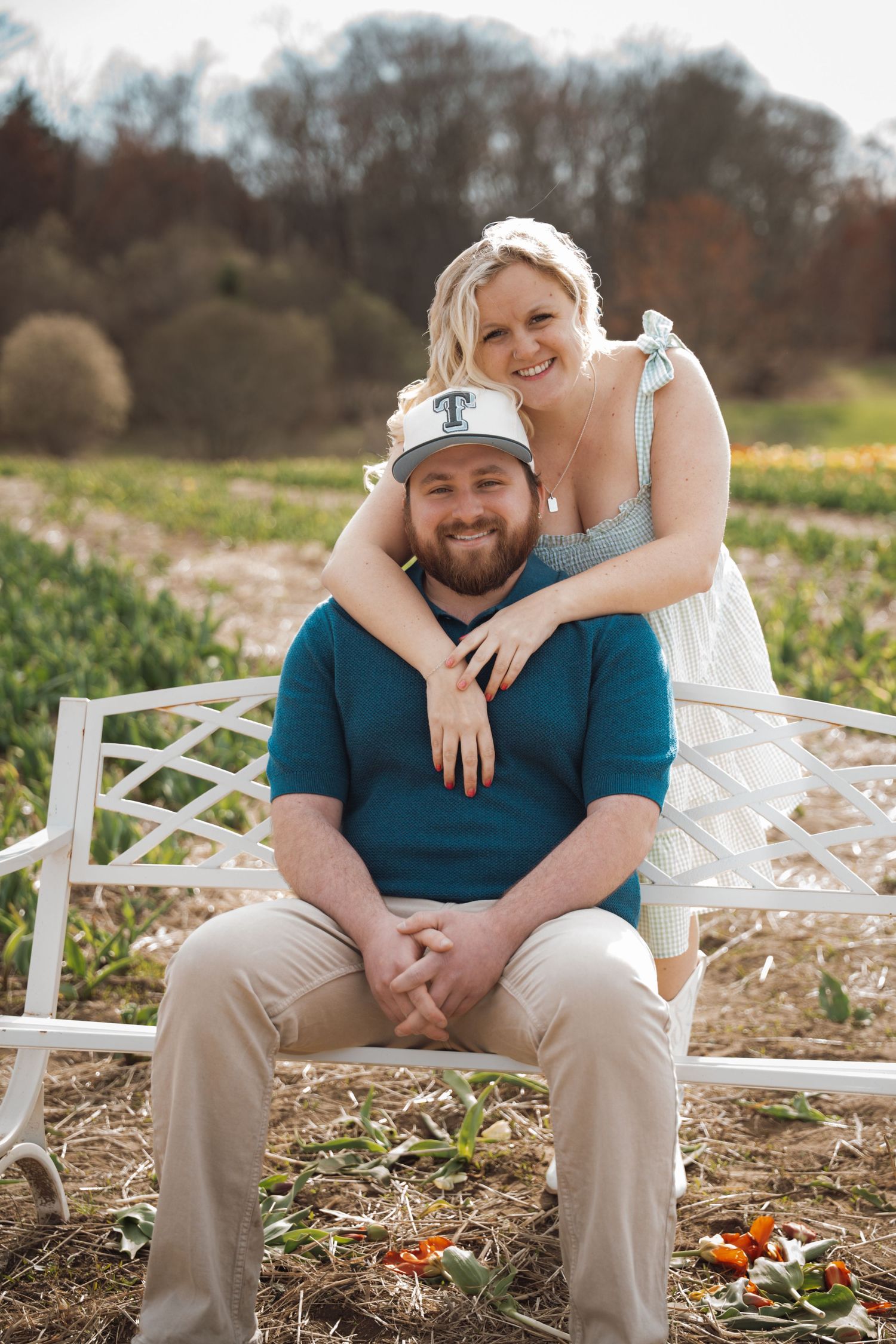 Couple posing on a white bench in a flower field, woman leaning over mans shoulders smiling.