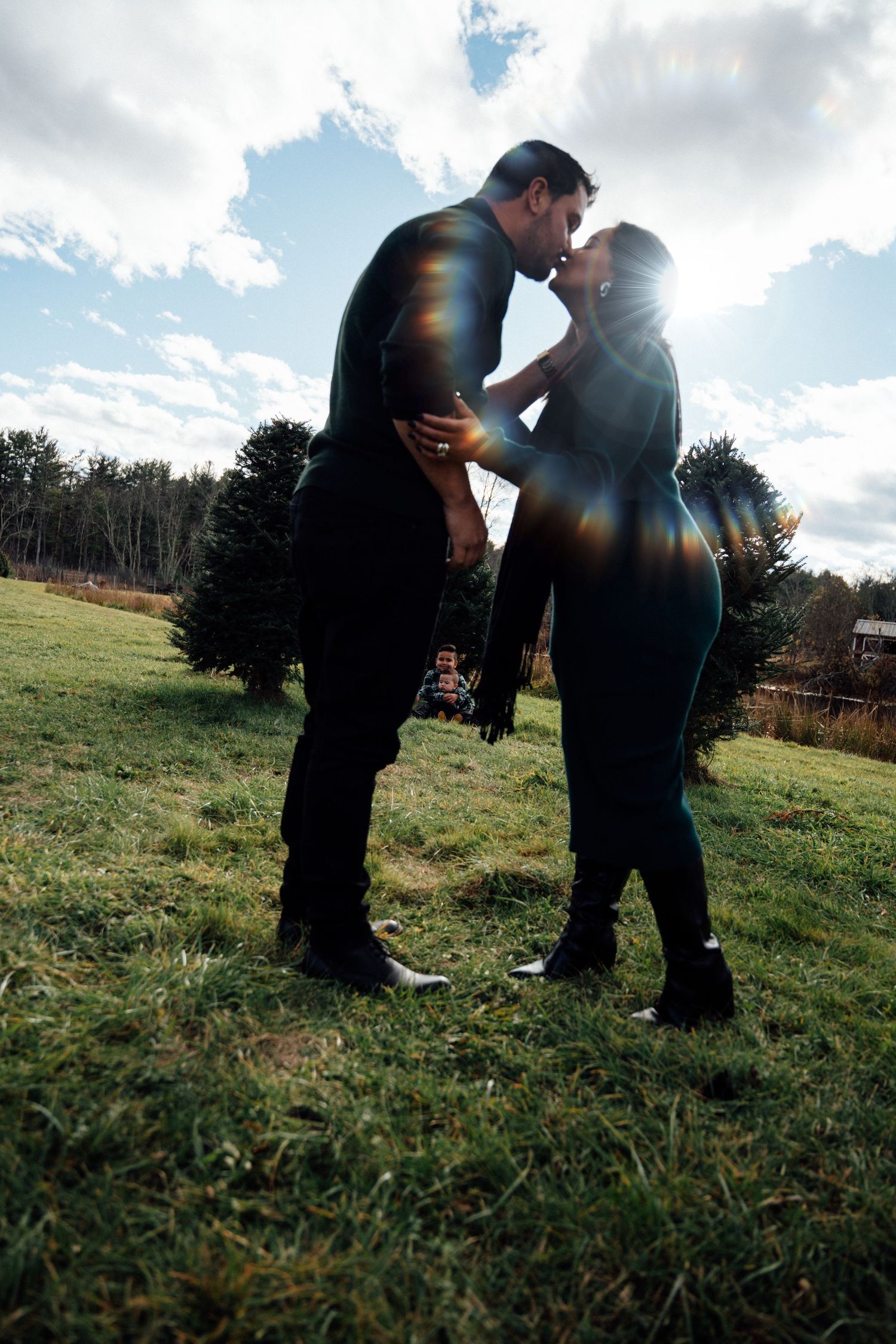 A romantic couple shares a kiss while standing in a sunlit field at a Christmas tree farm.