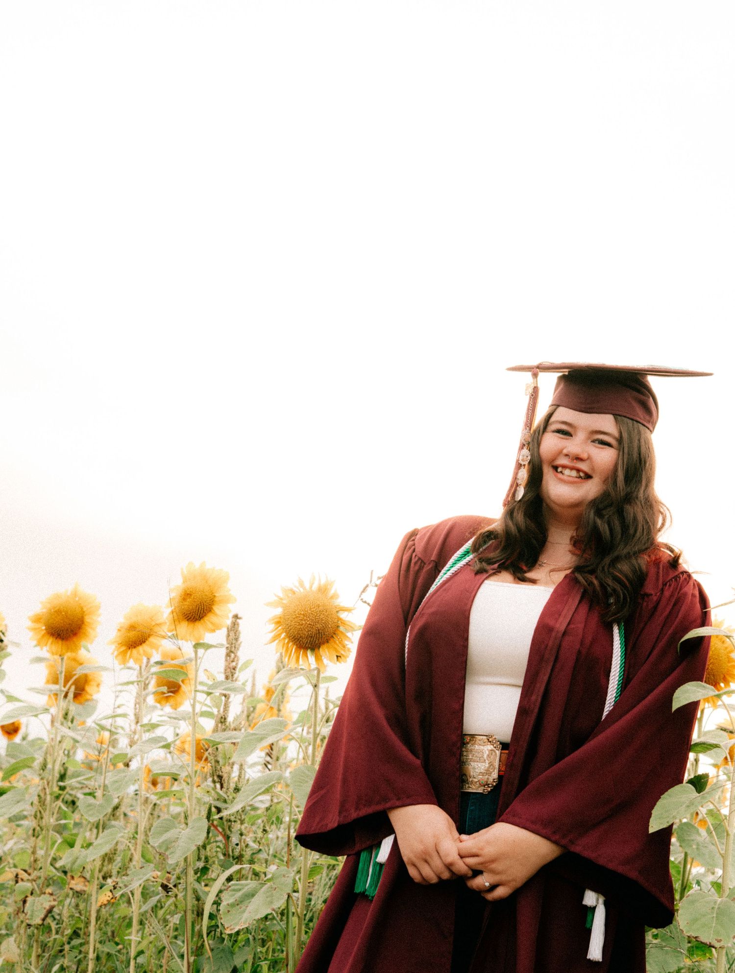 A graduate in maroon cap and gown smiles joyfully while standing in a sunflower field.