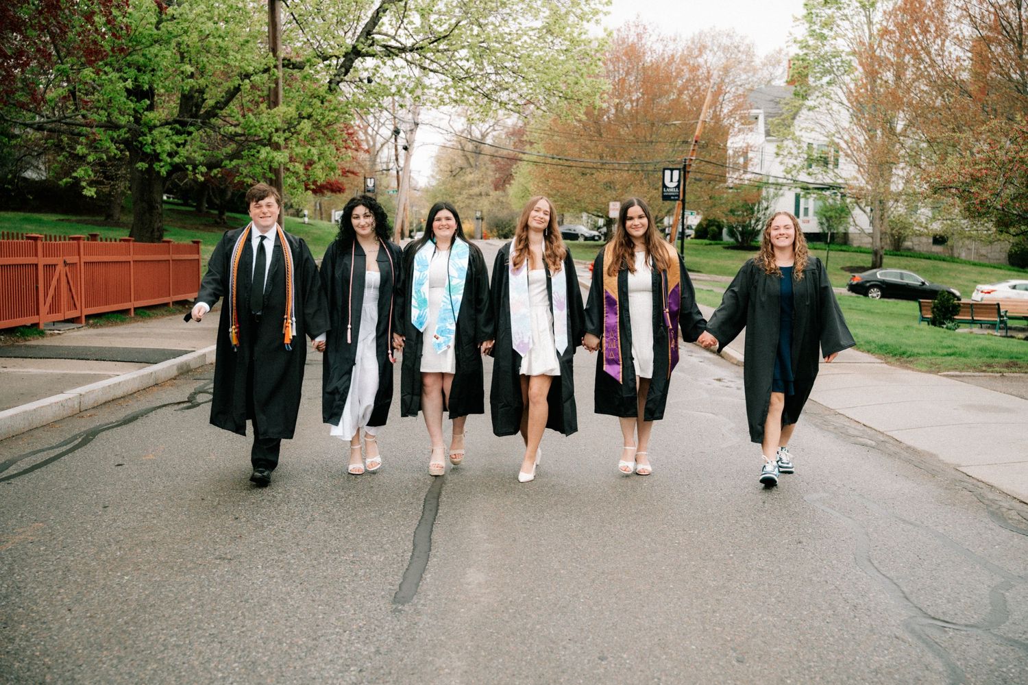 Seven graduates in black gowns walk hand-in-hand down a campus street surrounded by spring trees.