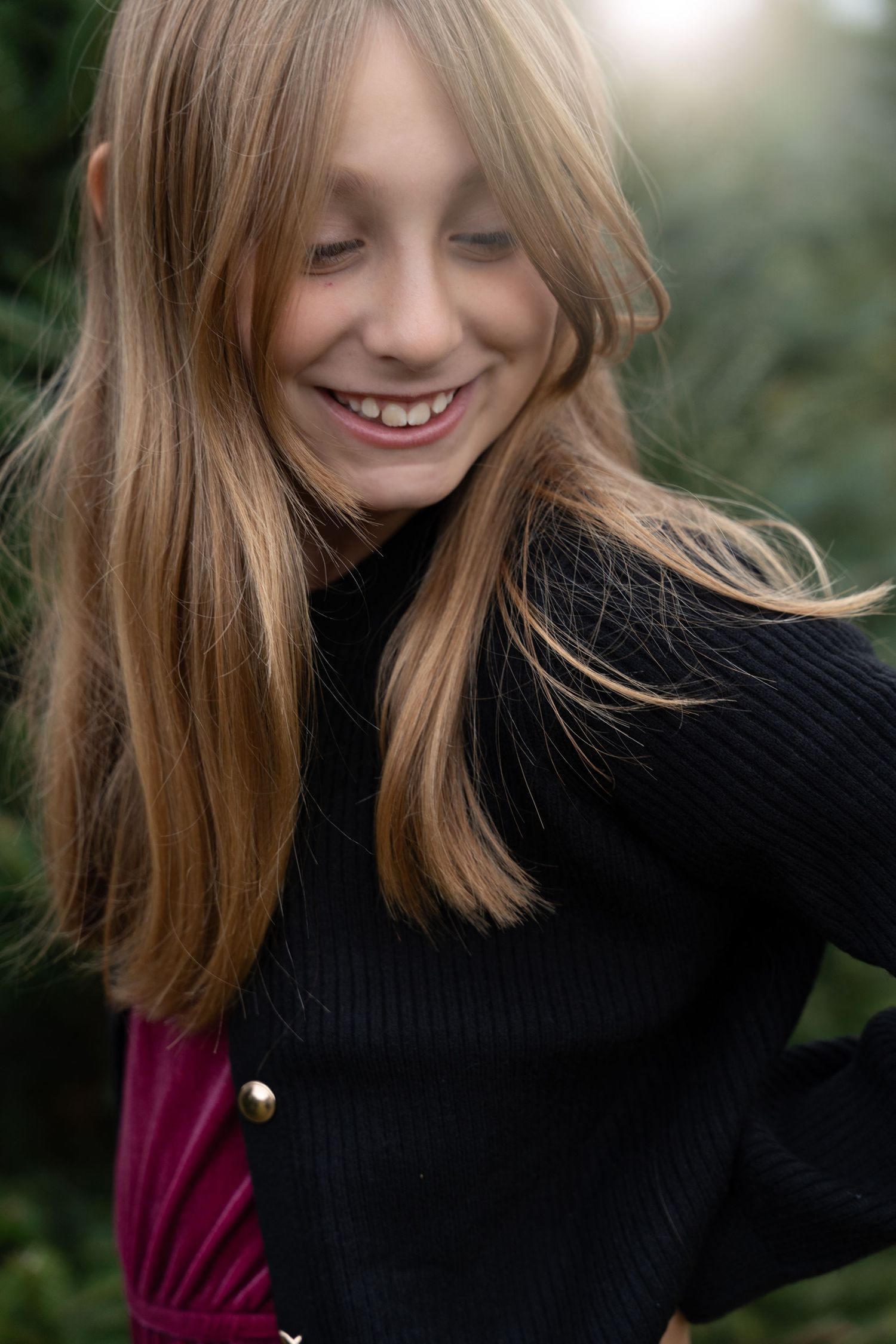 A young girl with long blonde hair laughs joyfully while standing among Christmas trees outdoors.