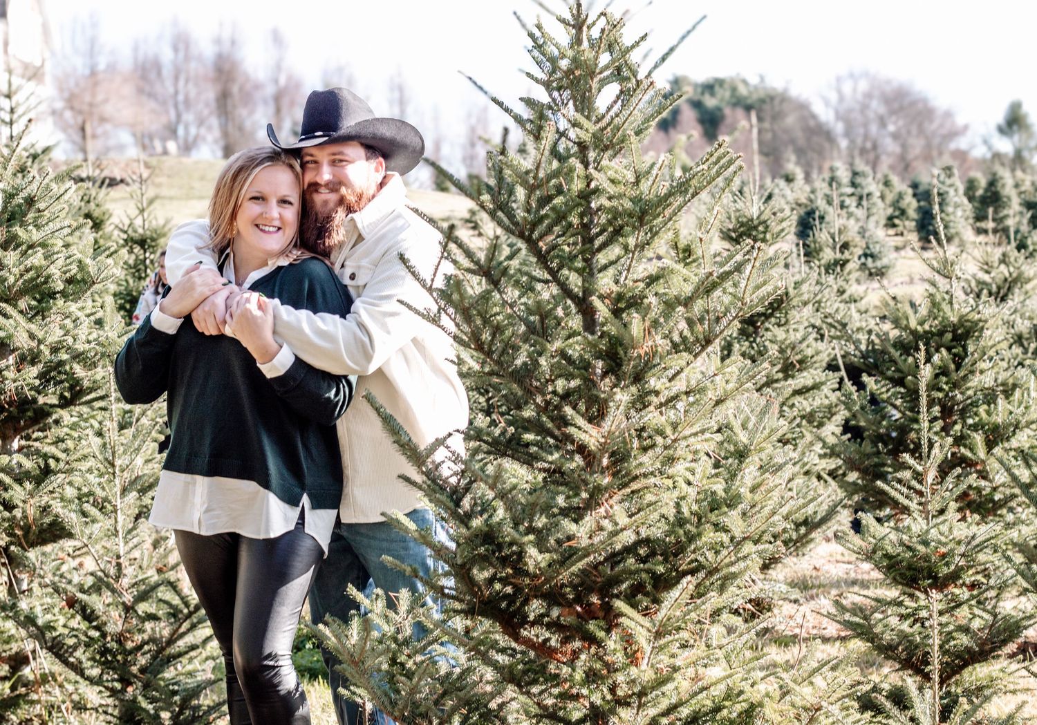 Smiling couple embracing and laughing together among rows of green Christmas trees at a tree farm.