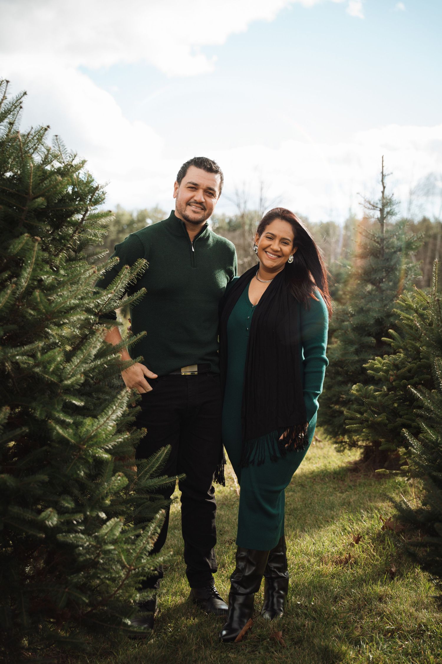 A smiling couple poses among green Christmas trees at a tree farm, both wearing coordinating dark green outfits.