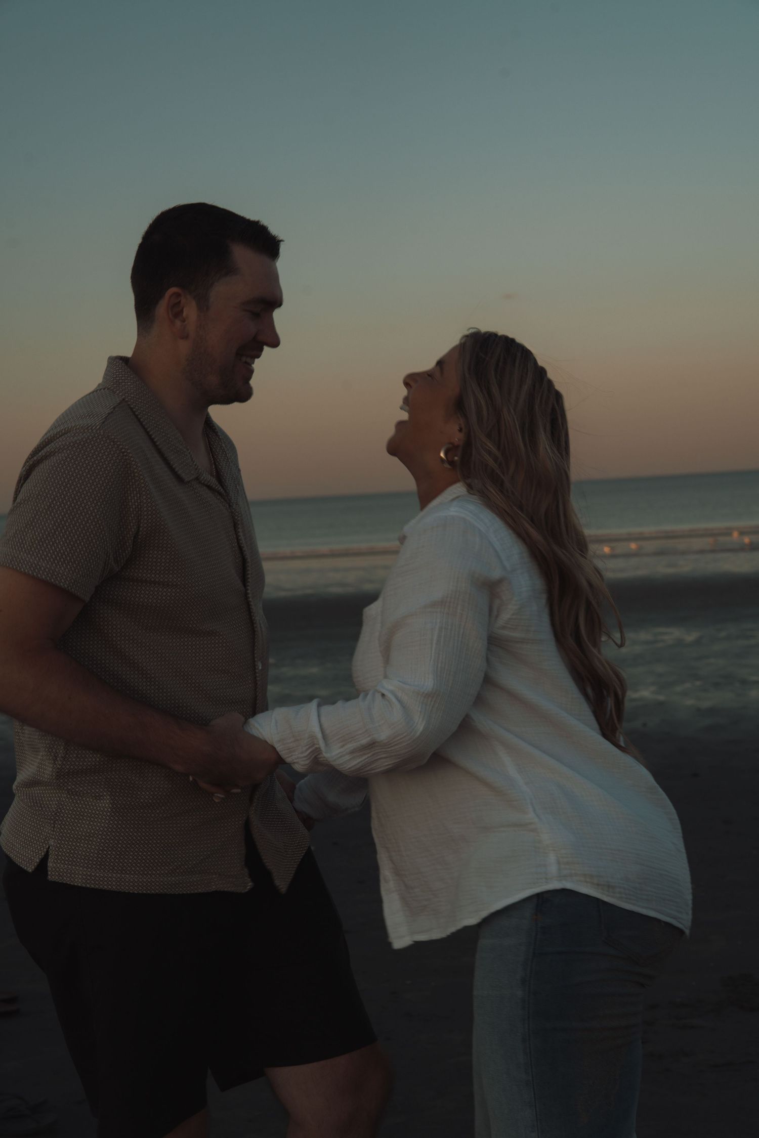 Couple holding hands and laughing together on a beach at sunset, man in gray shirt and woman in white top.