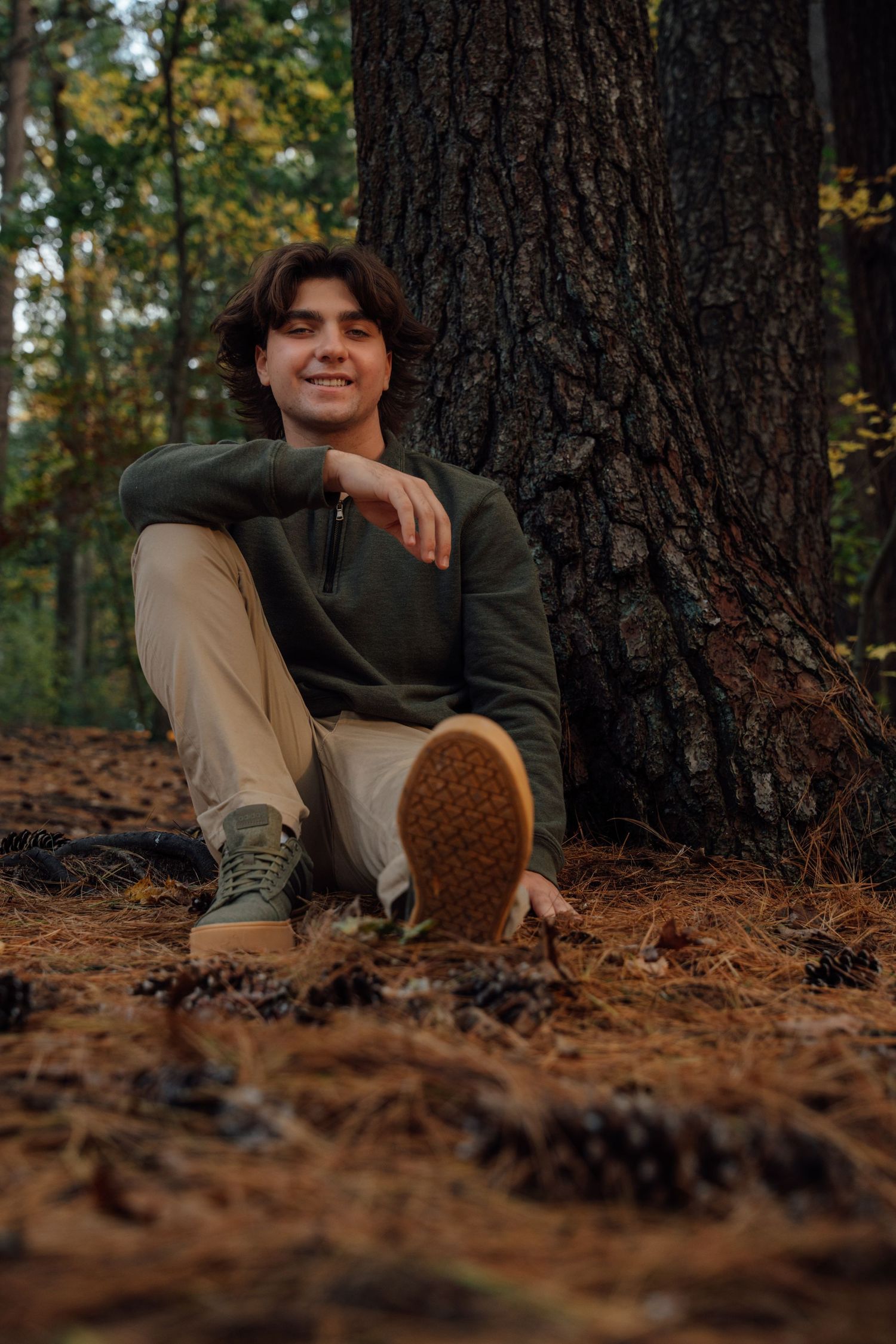 Smiling young man in green hoodie relaxes against large tree trunk in autumn forest.