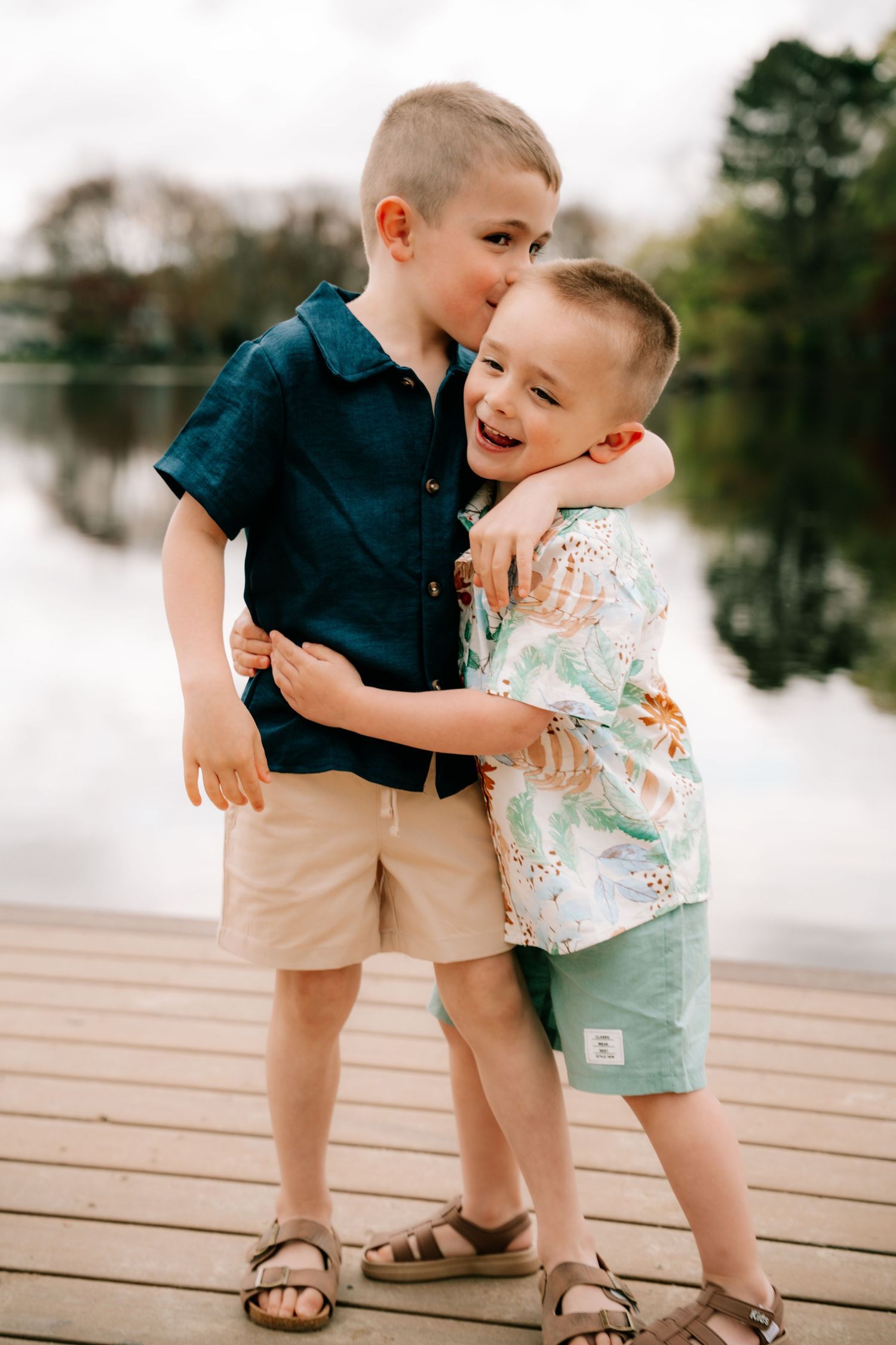Two young brothers hugging and laughing together on a wooden dock near a lake.