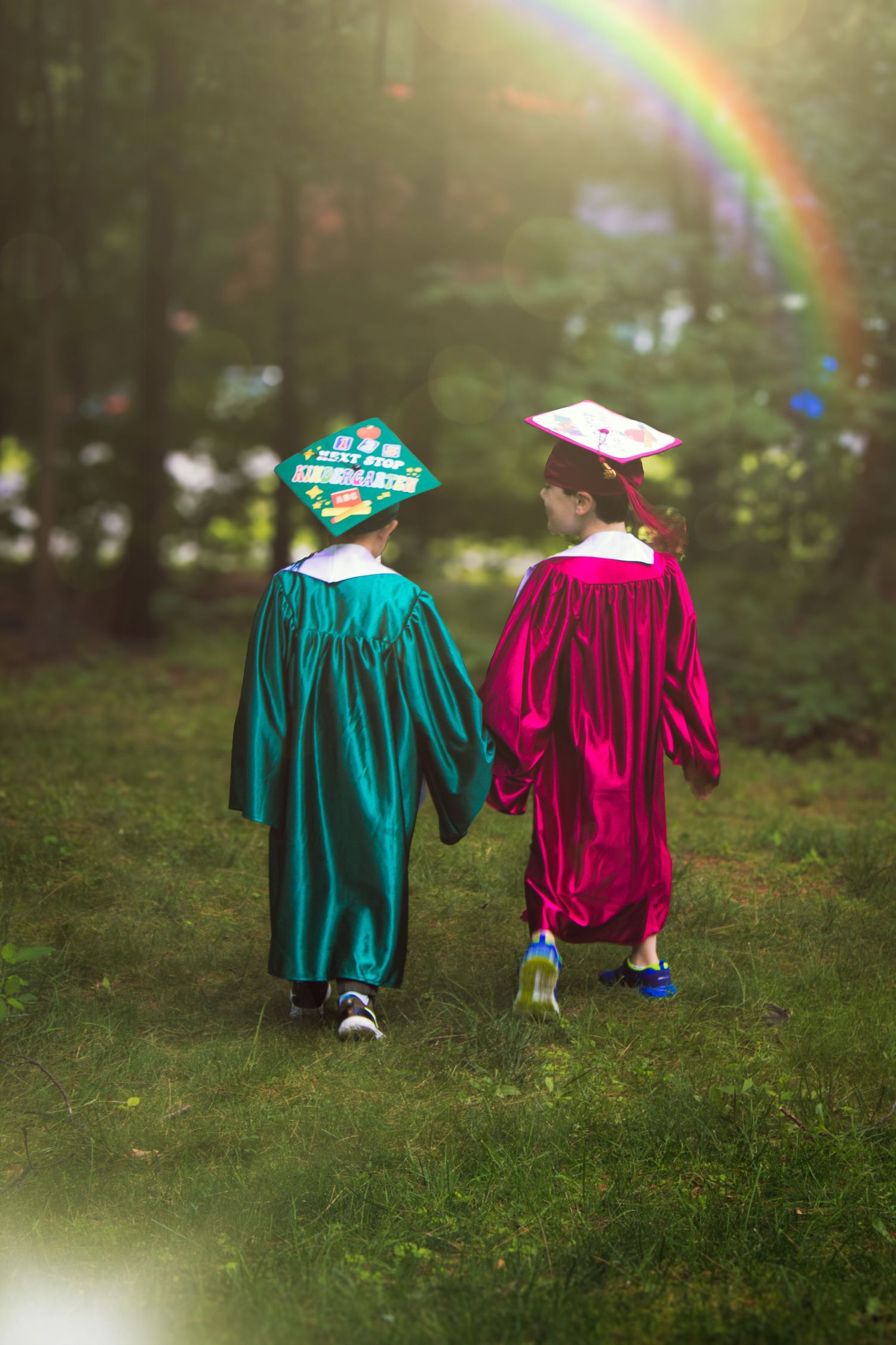 Two children in graduation gowns walking hand in hand with a rainbow arching behind them.