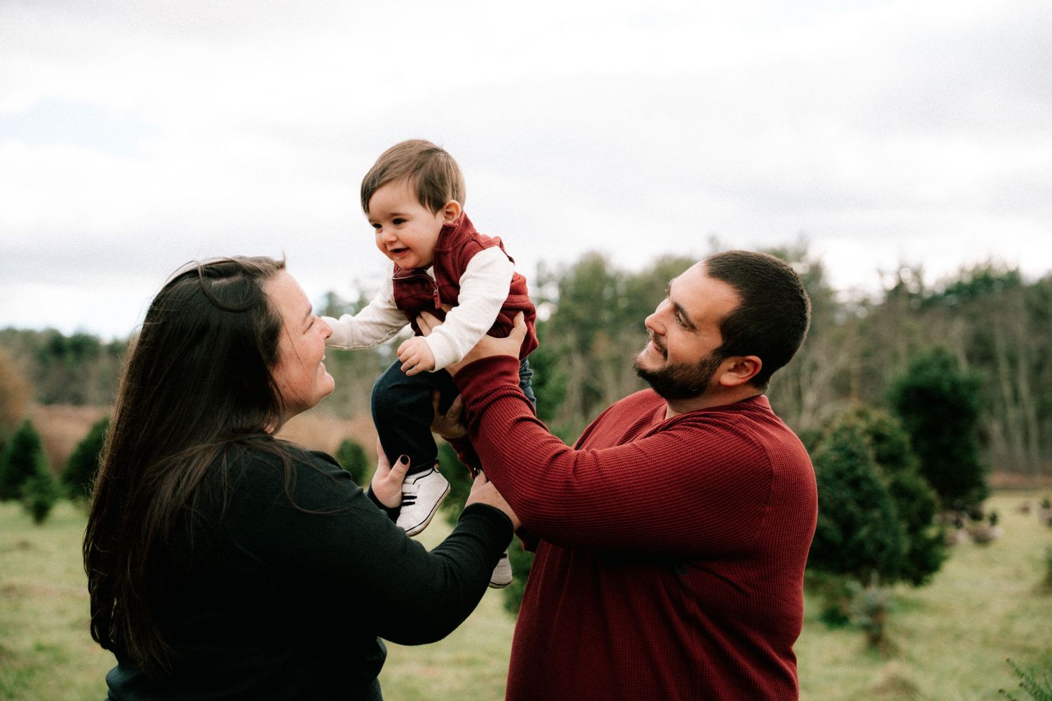 Family with baby at Christmas tree farm, parents lifting smiling toddler outdoors.