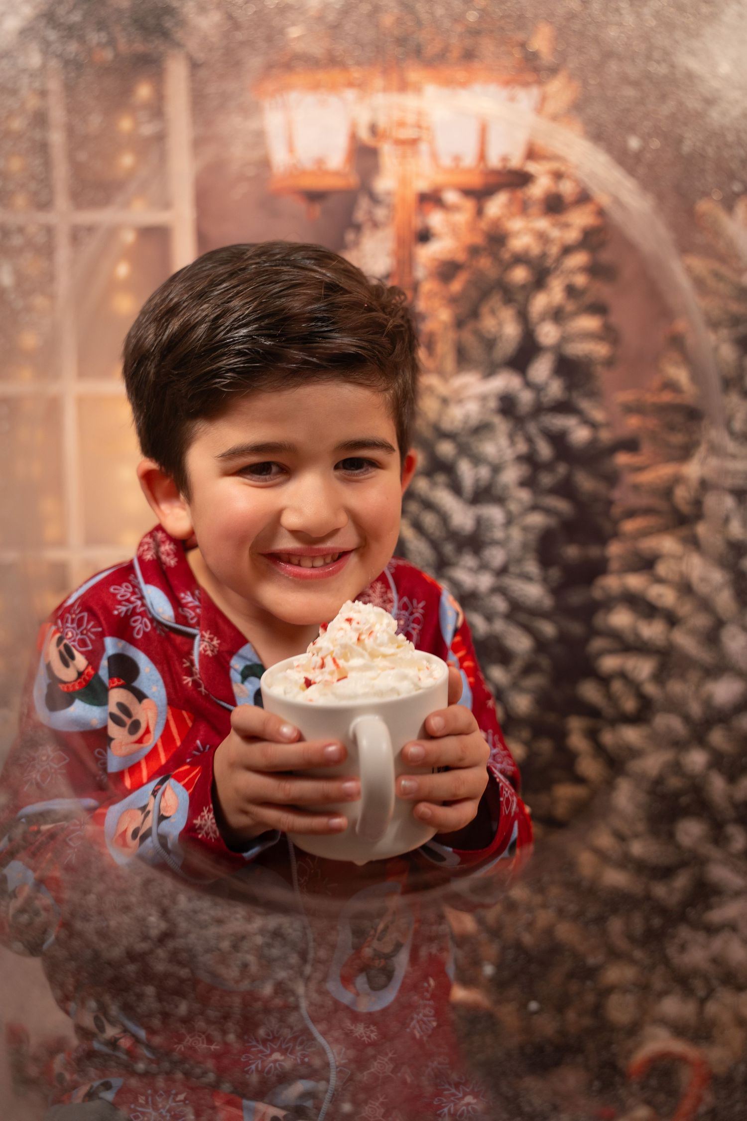 Smiling boy in Mickey pajamas holding hot cocoa with whipped cream.