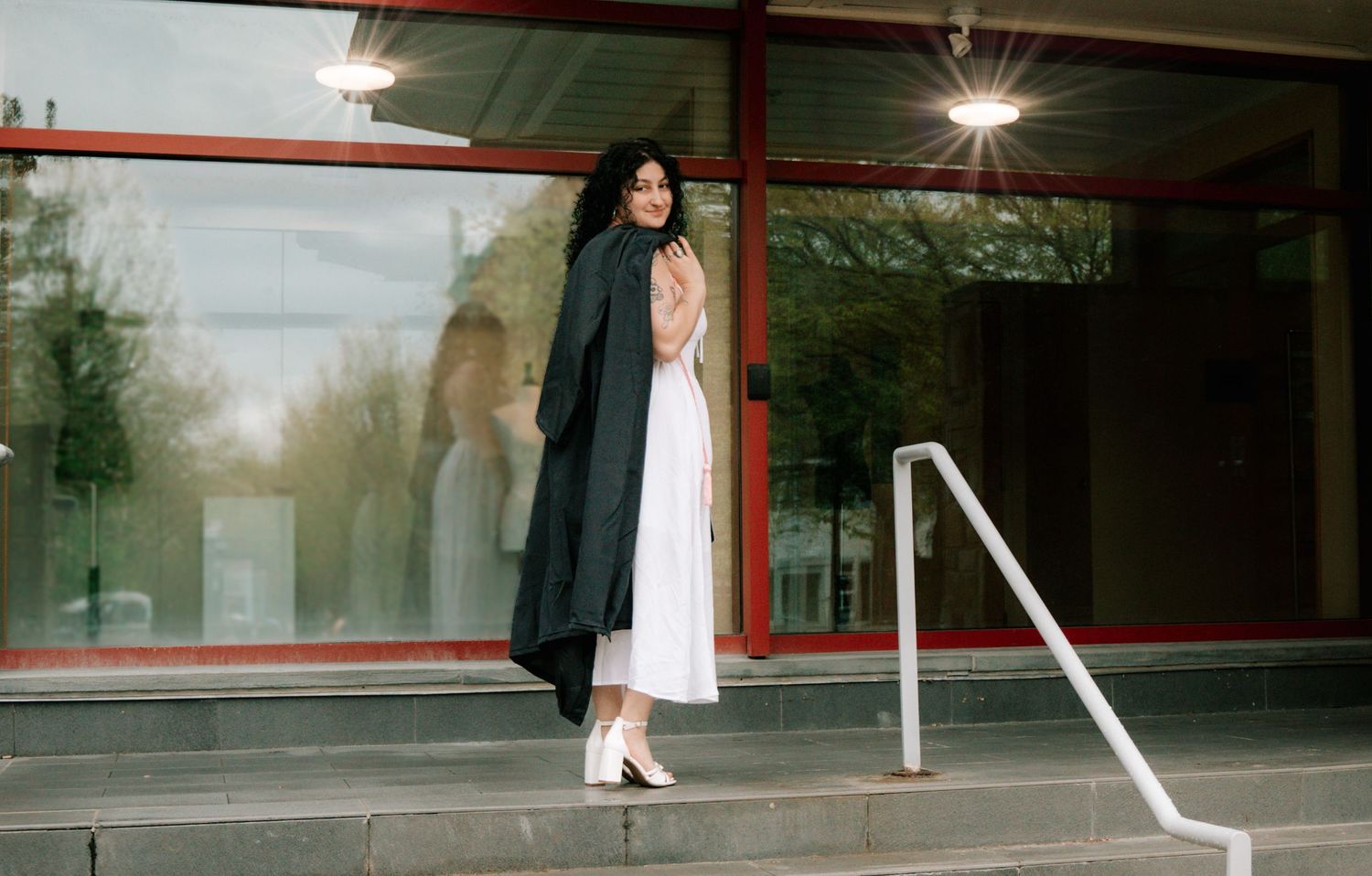 Female graduate in black robe and white dress posing on steps outside glass university building.