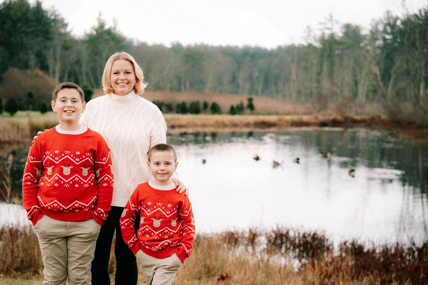 Mother and two sons in matching red holiday sweaters posing by a pond outdoors.