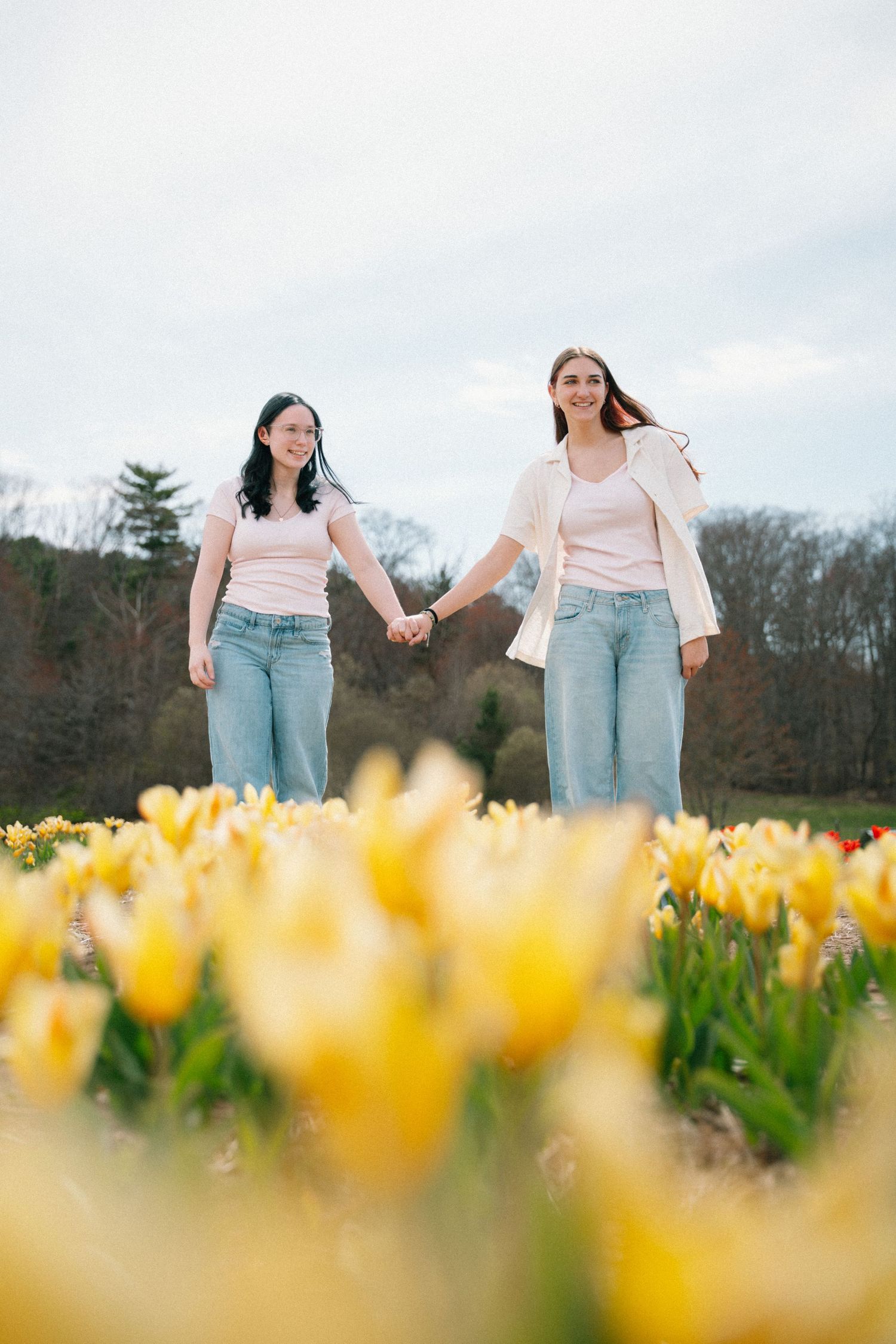 Two young women holding hands in a field of yellow tulips on a cloudy day.