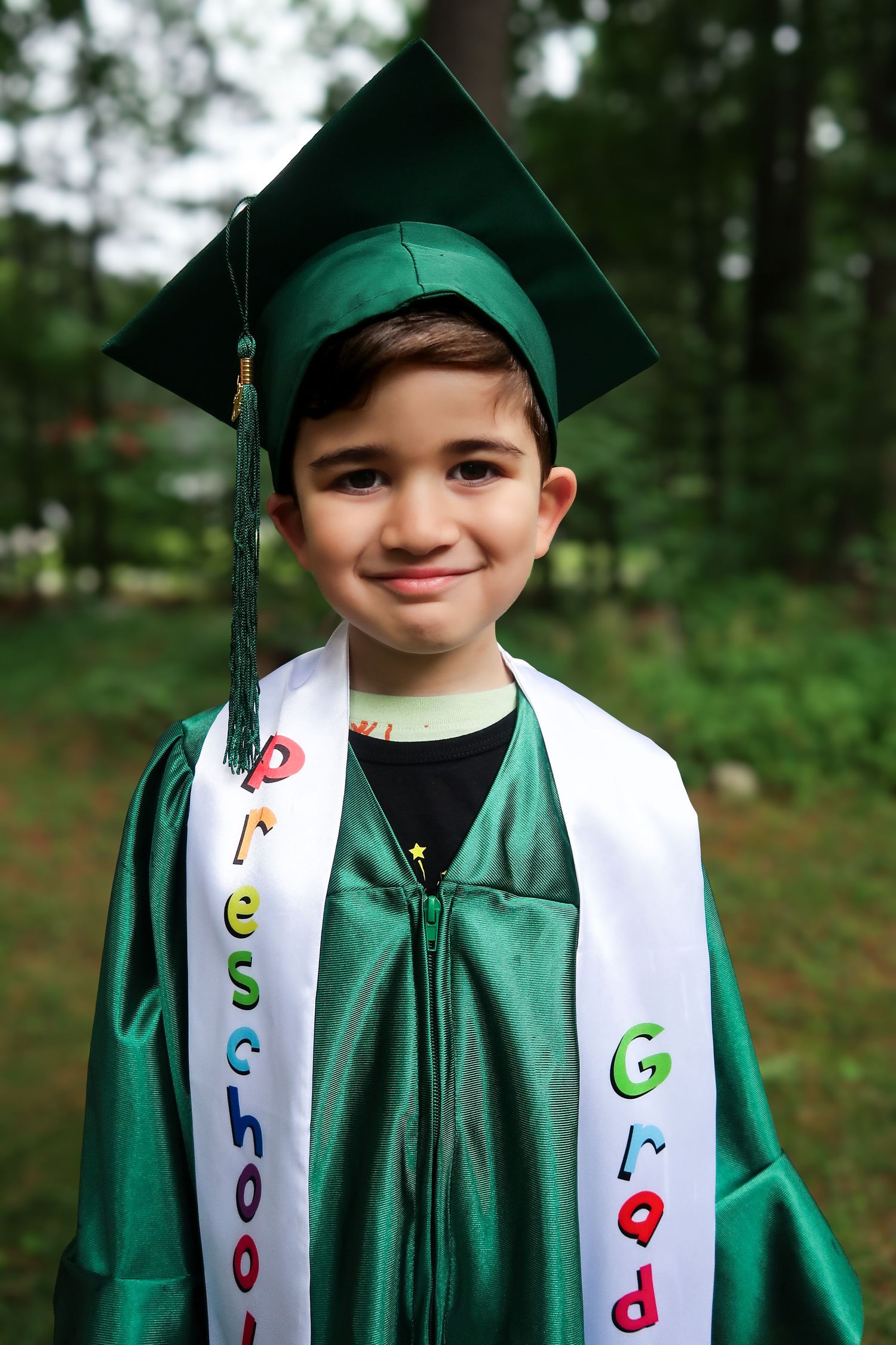 A young boy in a green preschool graduation cap and gown smiles wearing a graduate sash.