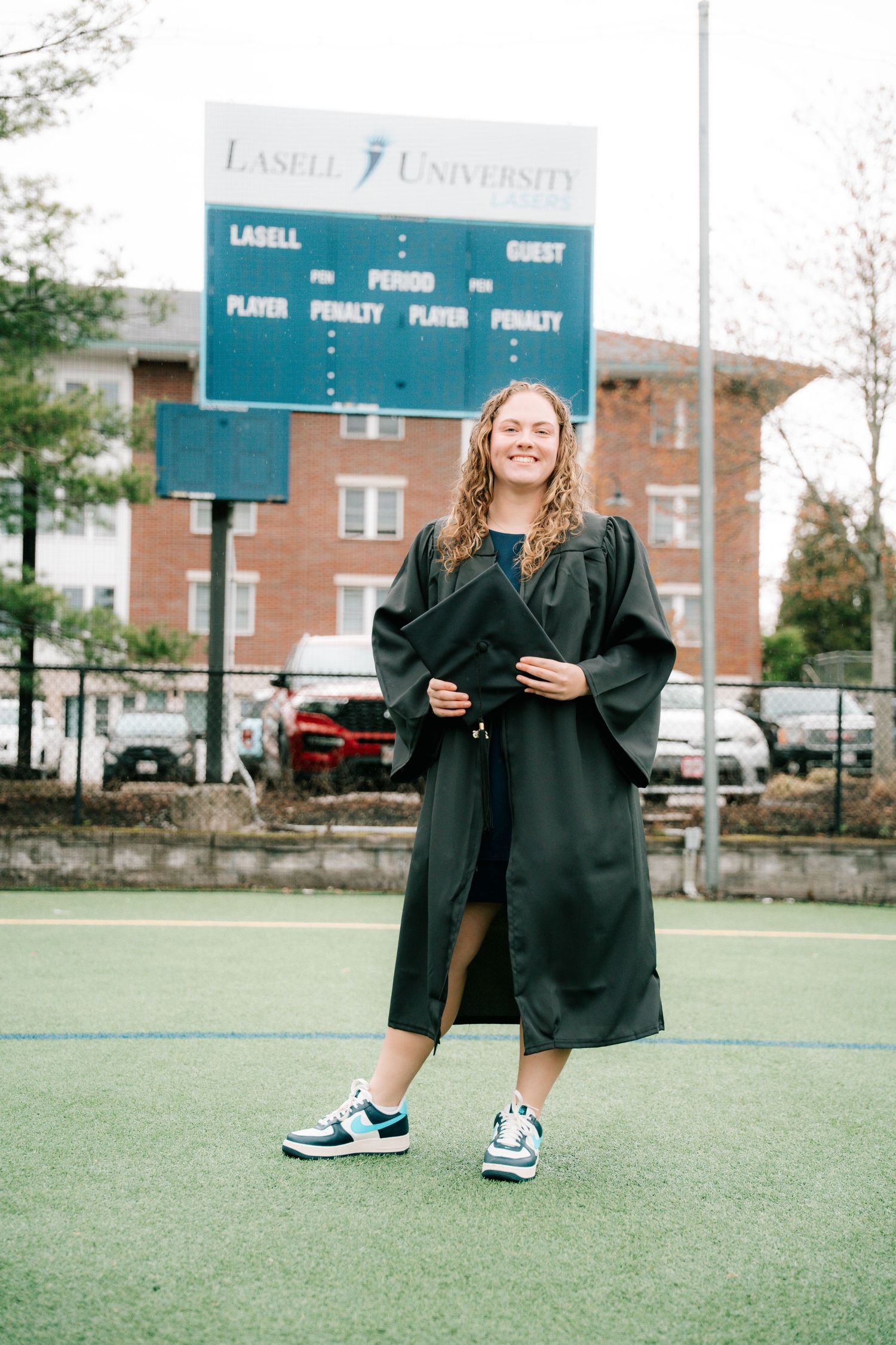 Smiling graduate in black robe standing on athletic field in front of Lasell University scoreboard.