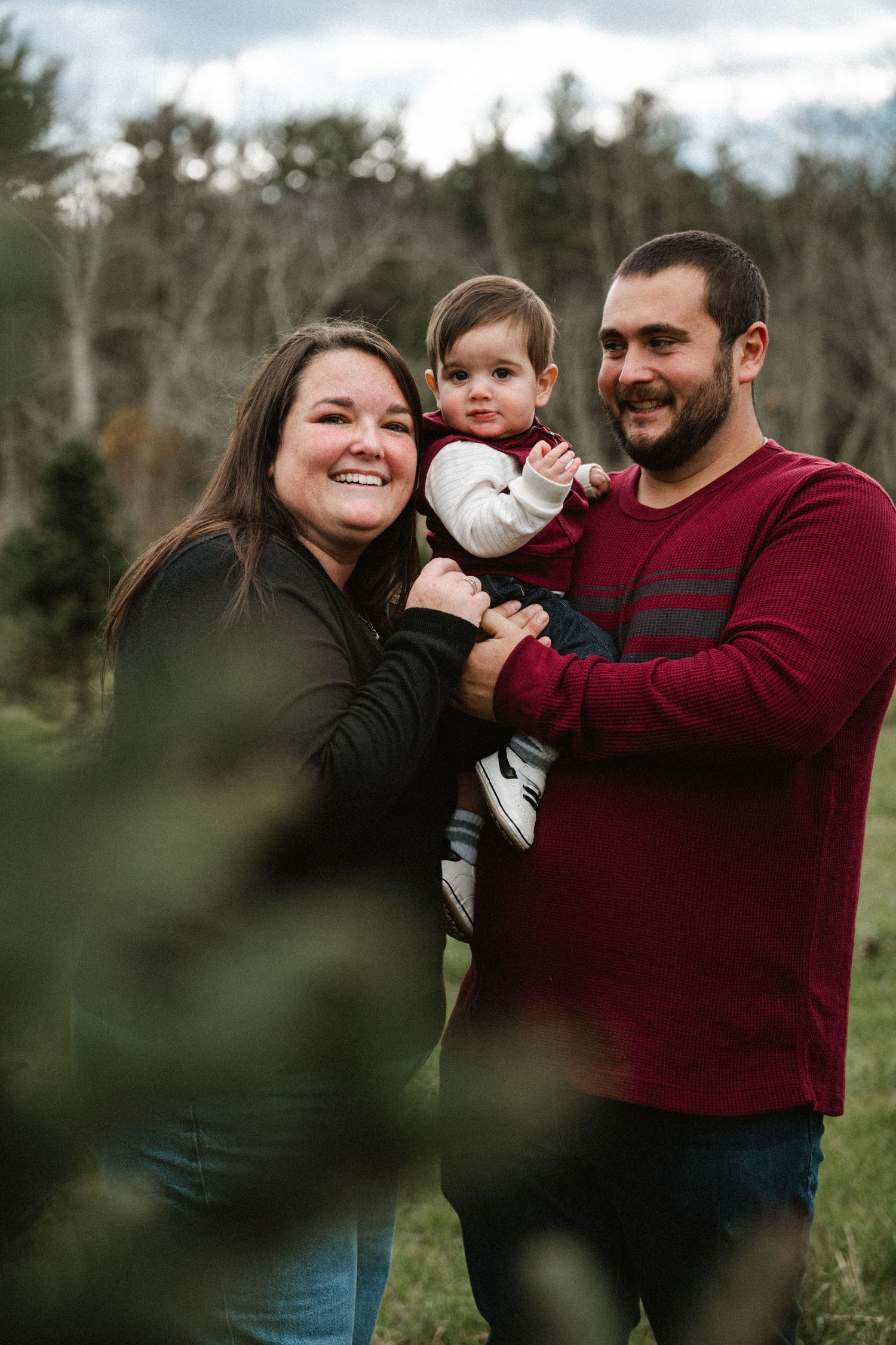 Smiling family of three with parents holding baby outdoors at a Christmas tree farm.