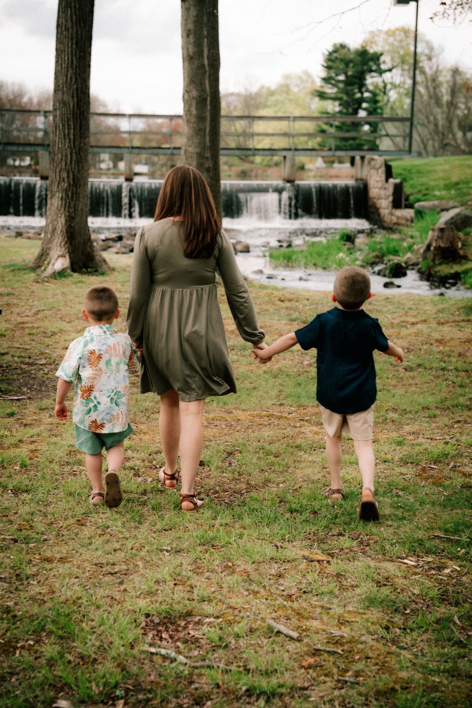 A mother walks hand-in-hand with her two young children through a lush green park near a scenic waterfall.