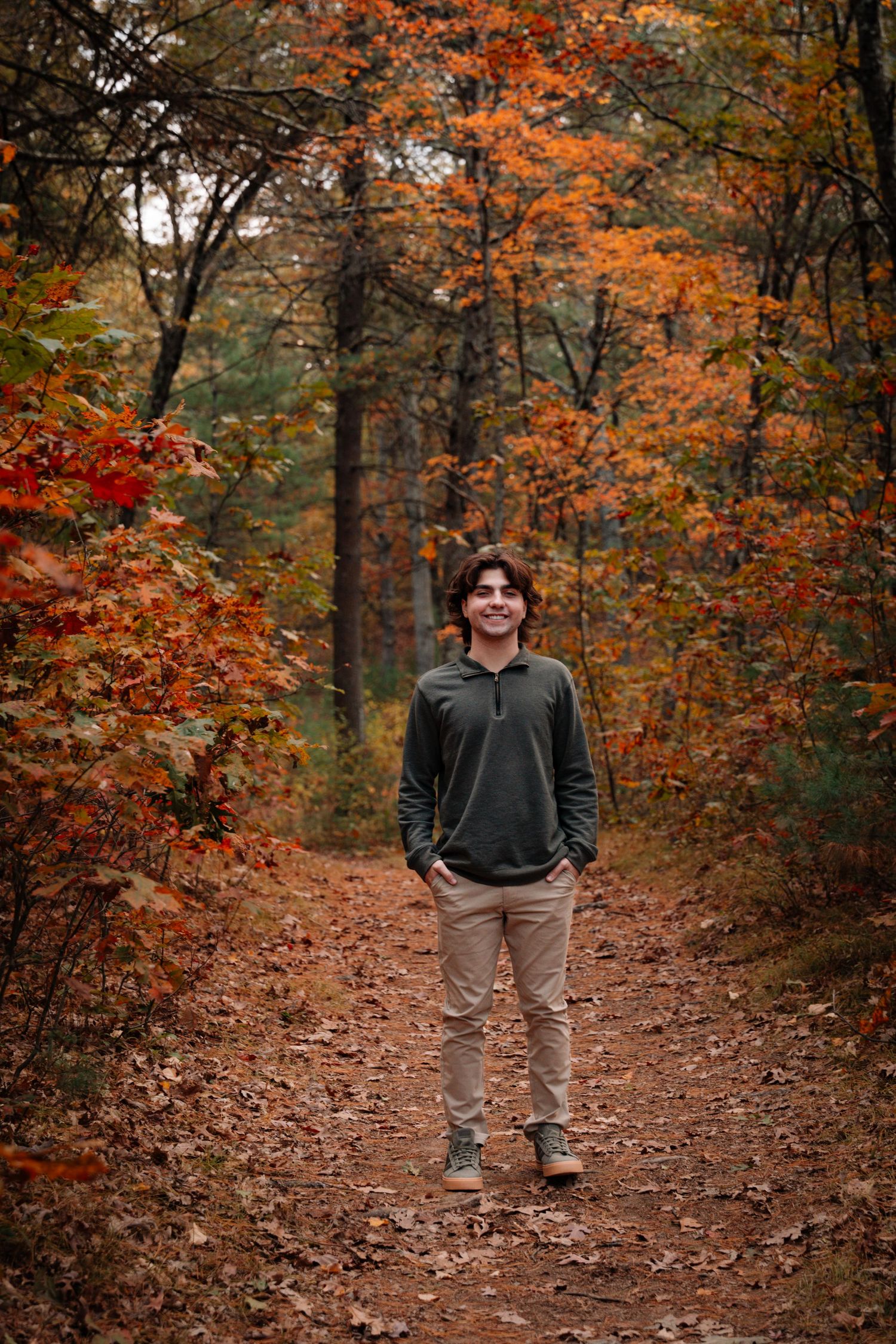 Smiling young man in green hoodie stands on leaf-covered path in colorful fall forest.