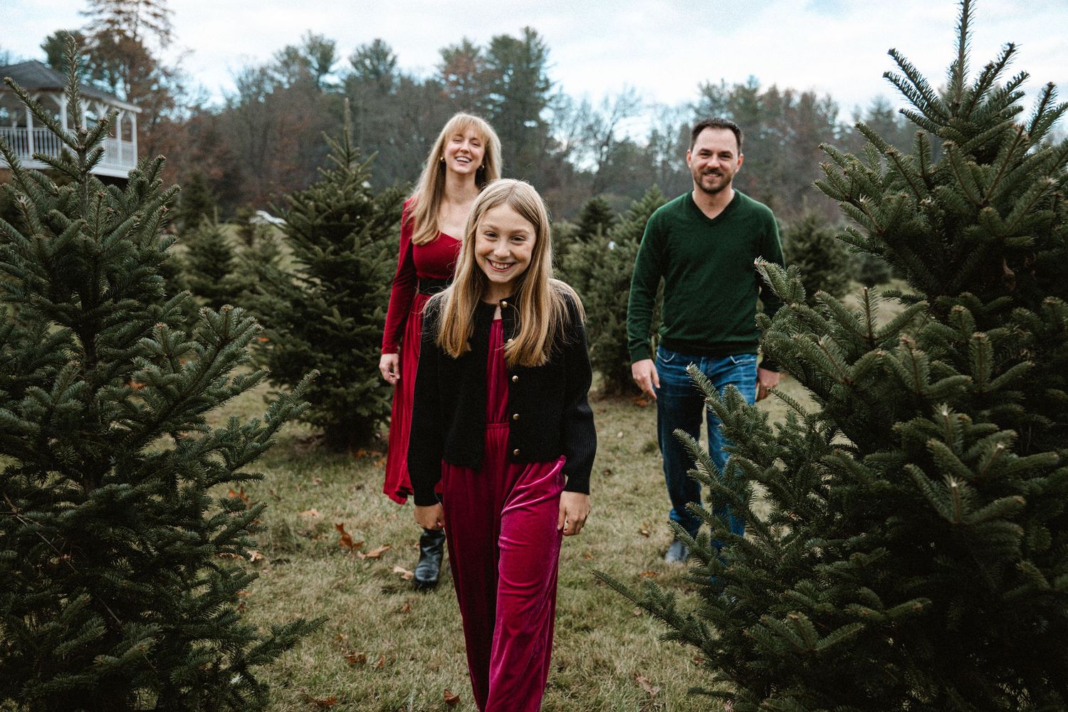 A cheerful family walks through rows of Christmas trees at an outdoor tree farm in holiday outfits.