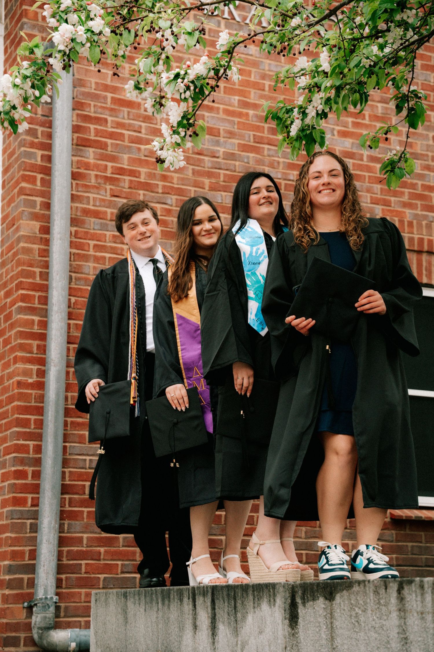 Four graduates in black gowns pose together on steps in front of a brick building with blooming trees.
