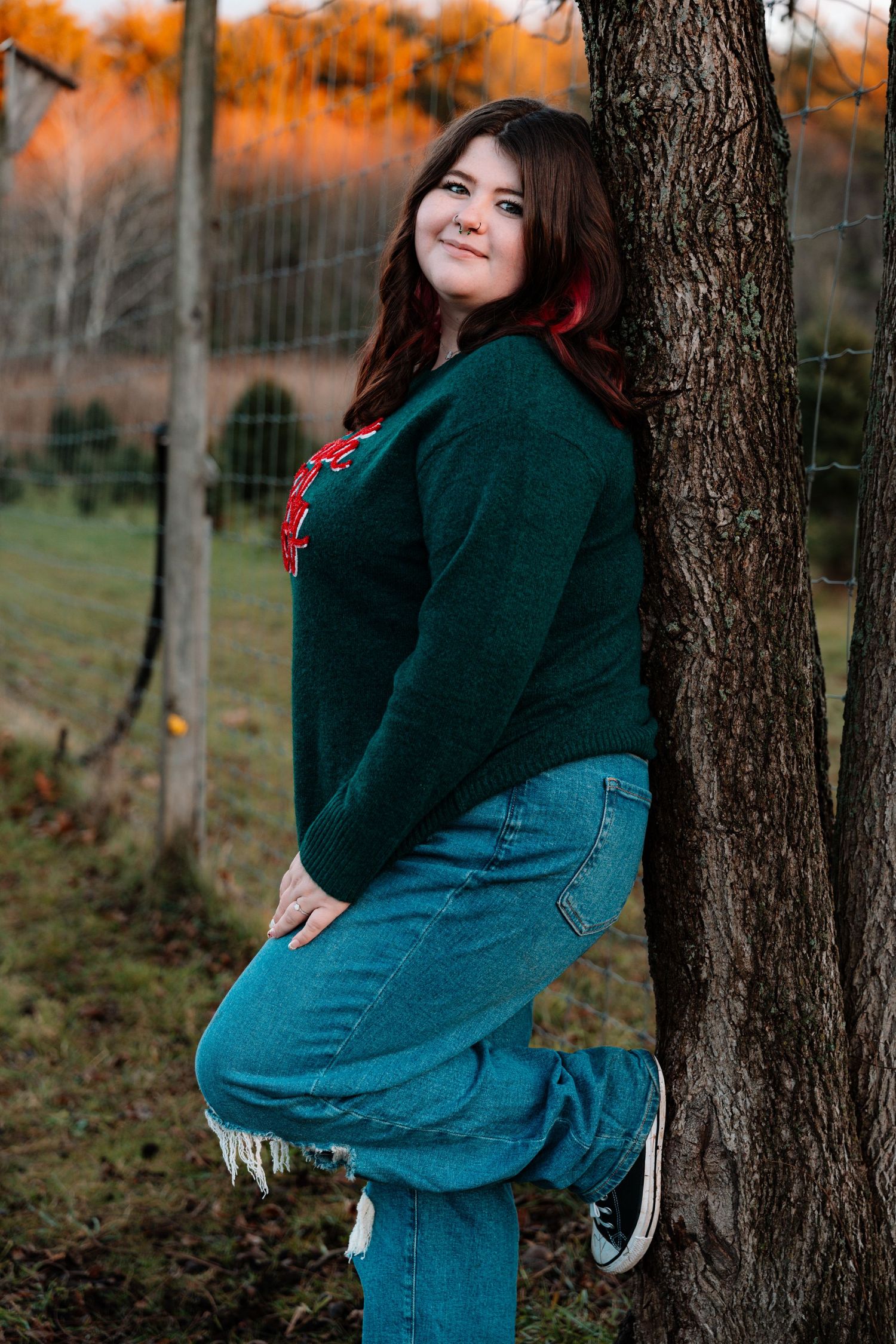 Smiling young woman in green sweater and jeans leaning against a tree at golden hour outdoors.