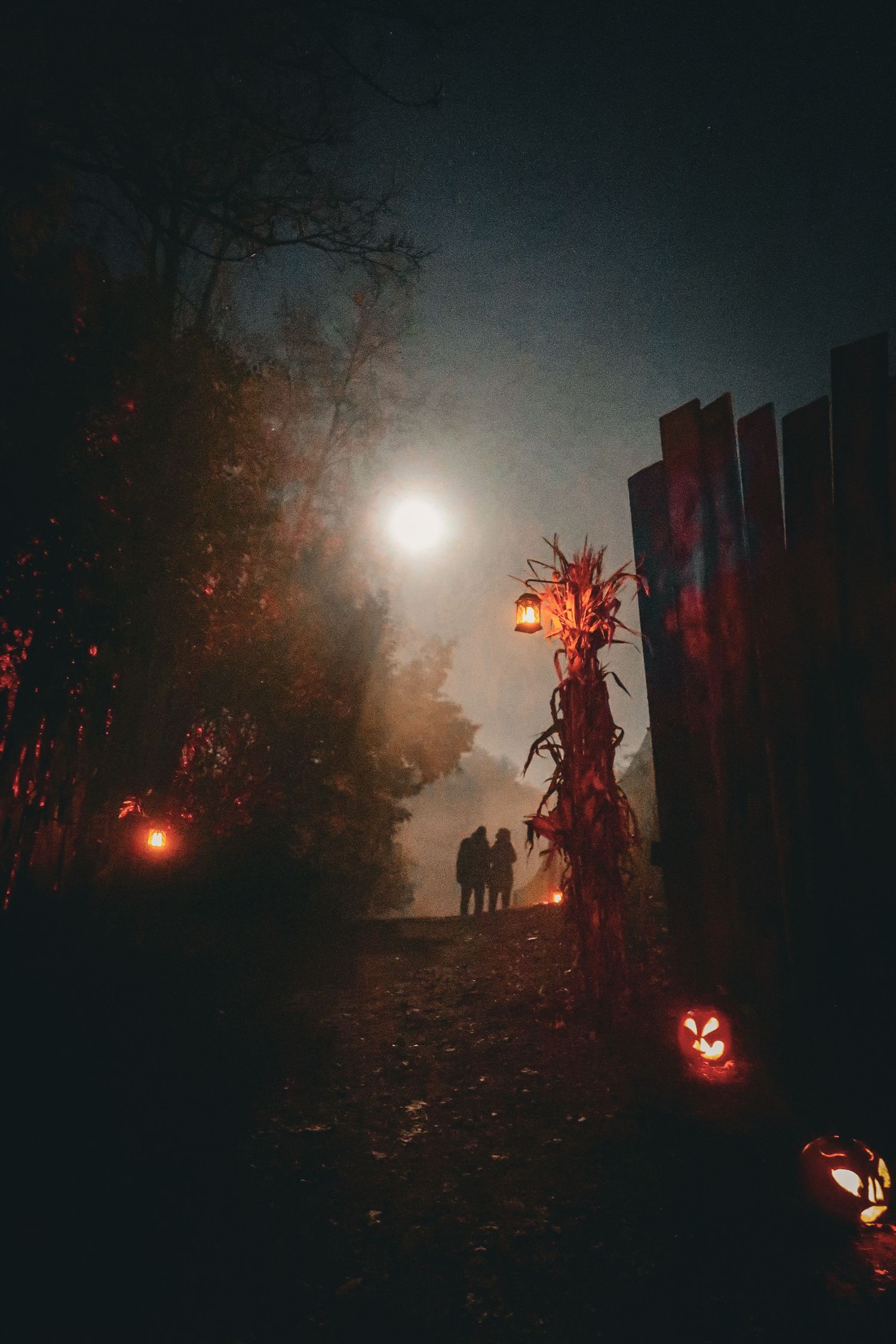Spooky Halloween night scene with jack-o-lanterns, fog, and silhouetted figures on a haunted path.