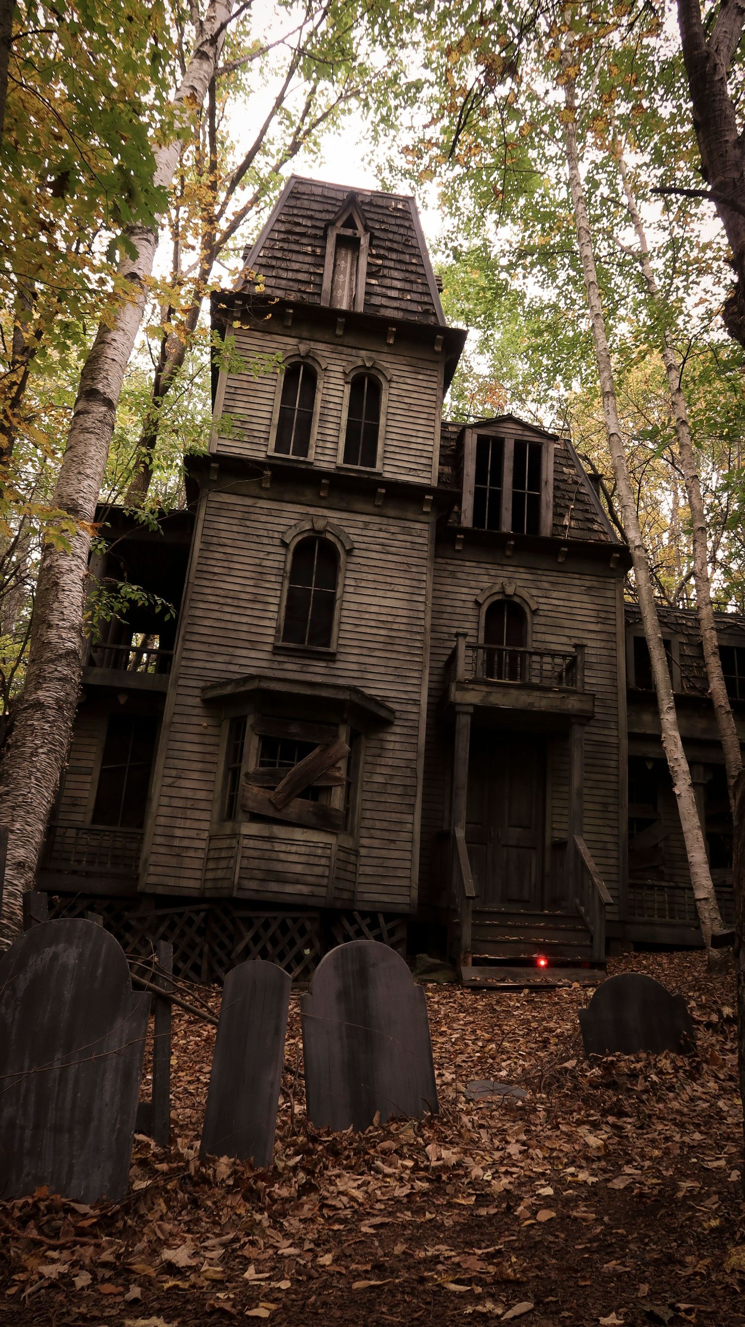 Abandoned haunted Victorian house with gothic windows surrounded by autumn trees and tombstones.