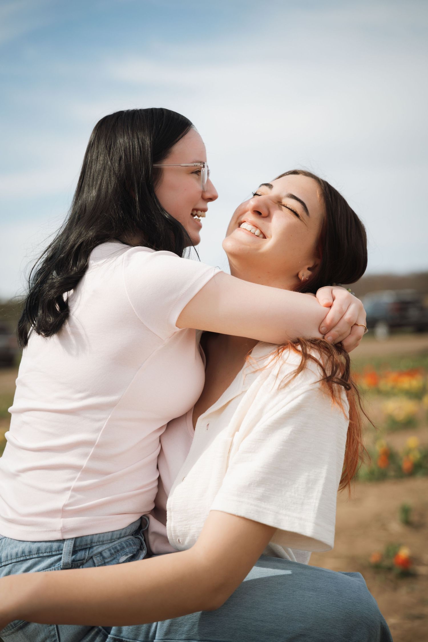 Two young women laughing and embracing outdoors in a flower field under a blue sky.