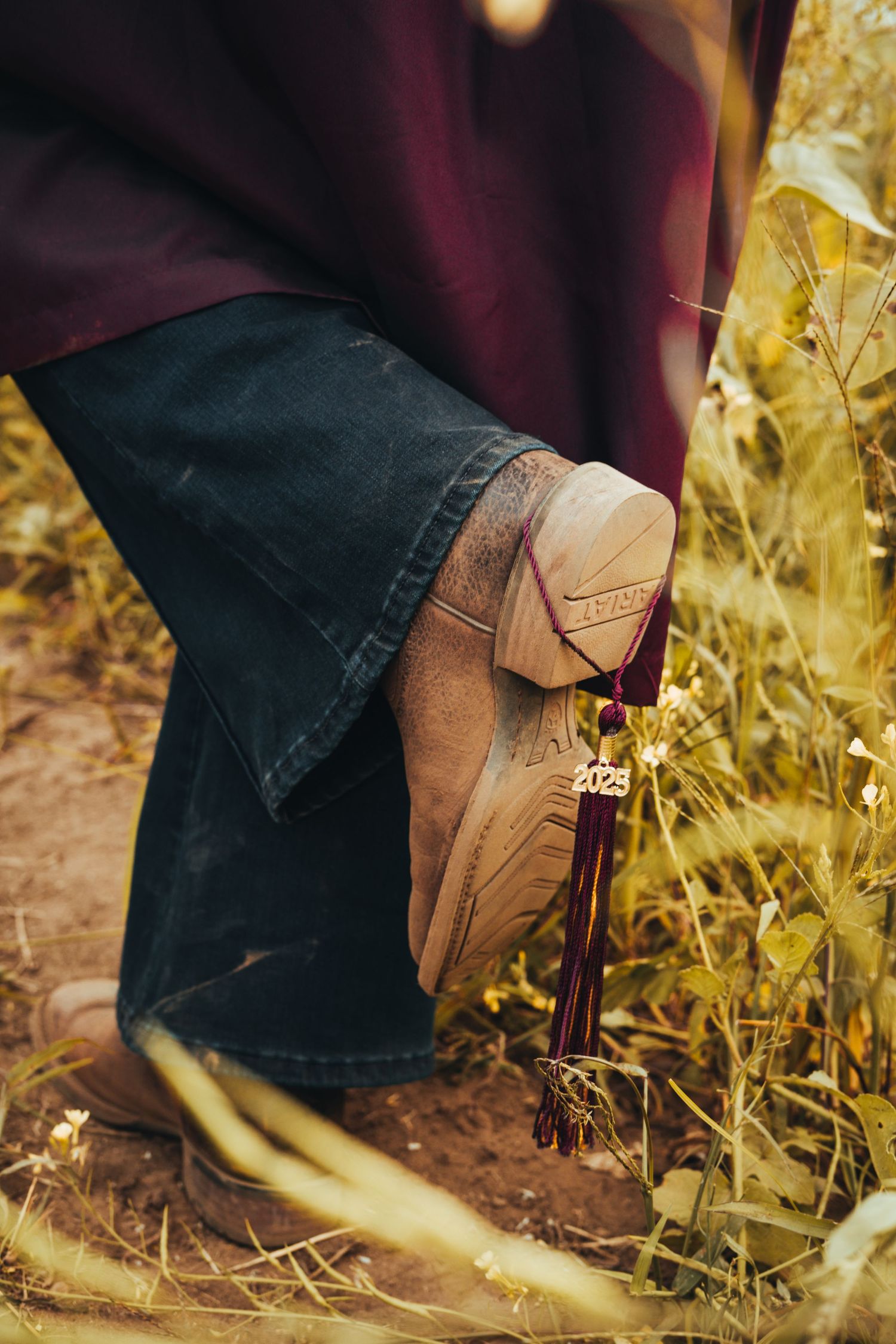 Close-up of a graduation tassel with 2025 charm on a tan boot in a golden field.
