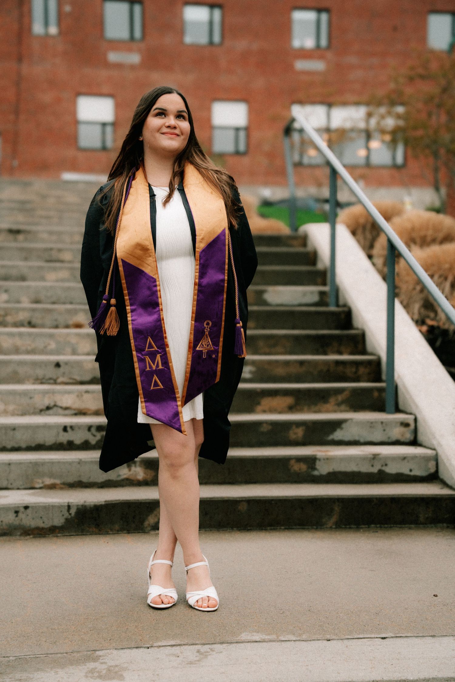 A smiling graduate in black gown wearing a purple and gold Delta sorority stole stands on campus steps.