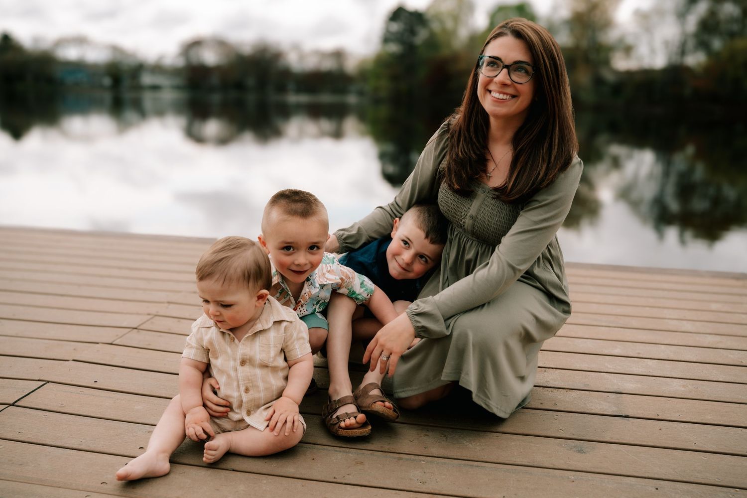 Smiling mother sitting on a wooden dock with her three young children by a reflective lake.