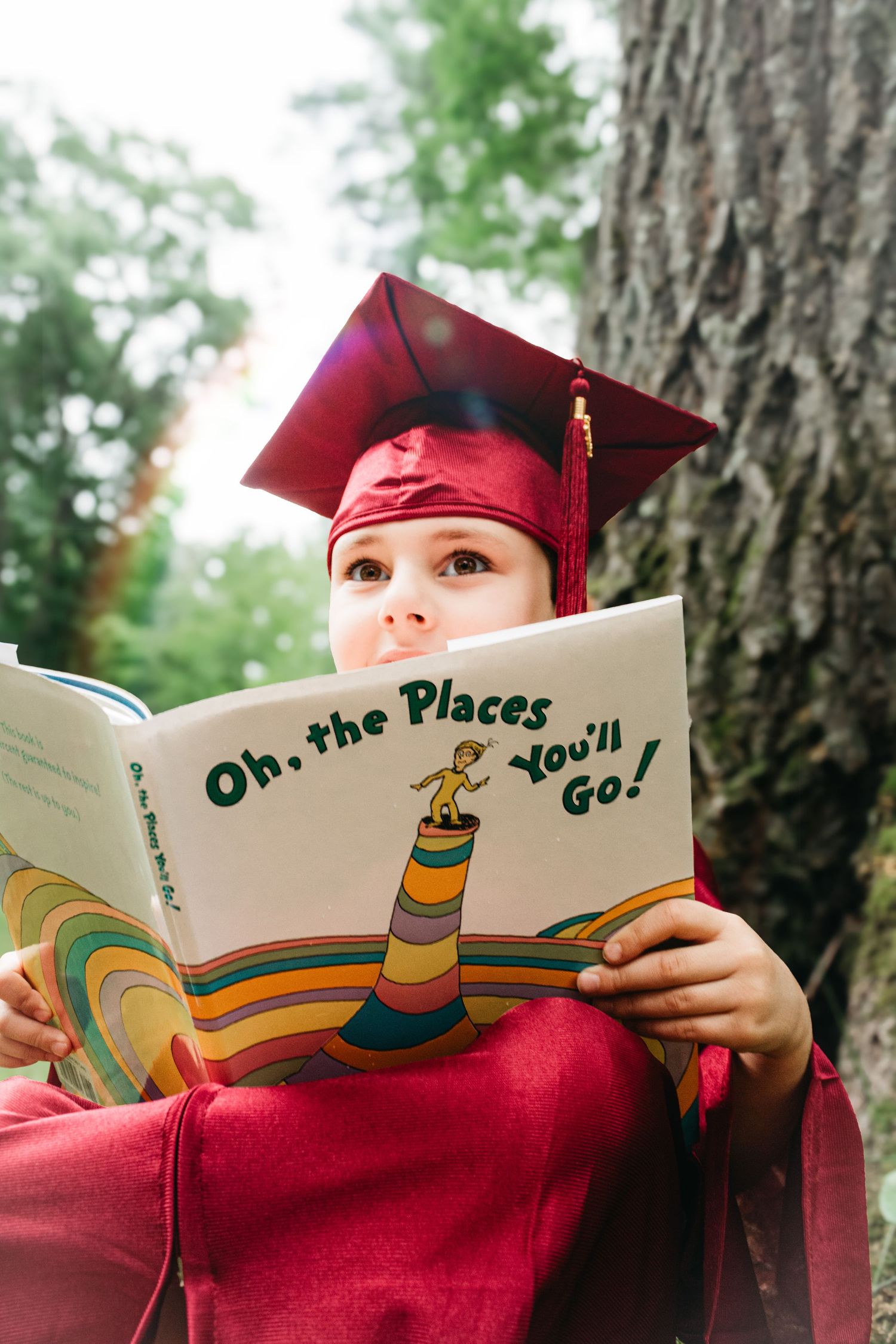 A young child in a red graduation cap reads Dr. Seuss Oh the Places Youll Go outdoors.
