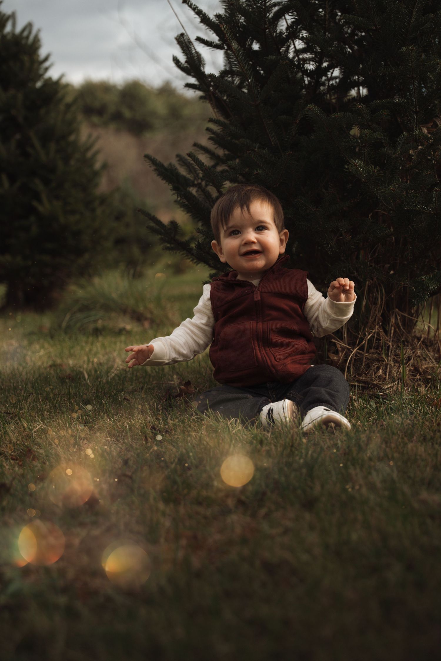Smiling baby in burgundy vest sitting on grass beside a pine tree outdoors.