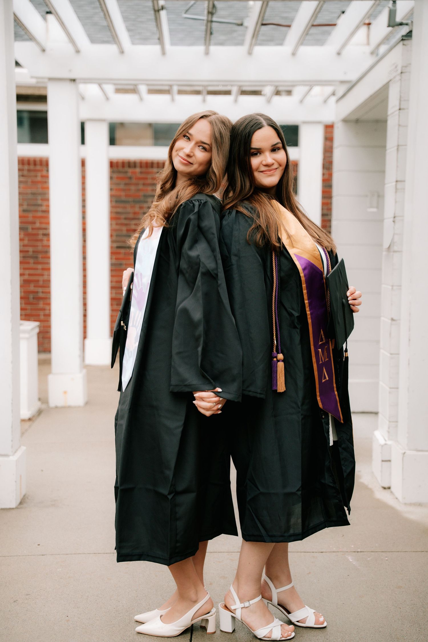 Two female graduates in black robes posing back-to-back under a white pergola.