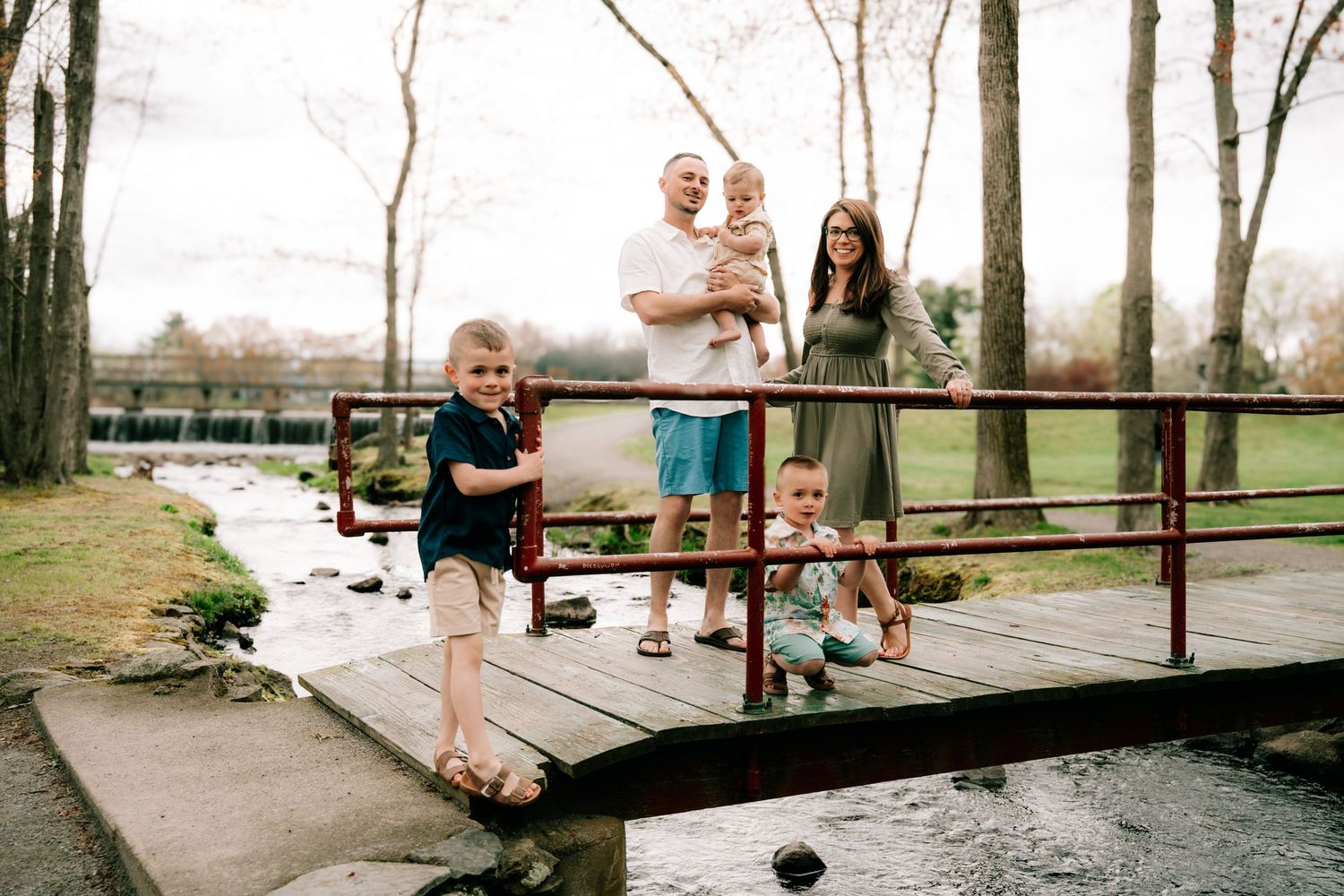 Happy family of five standing on a small wooden bridge over a stream in a park.