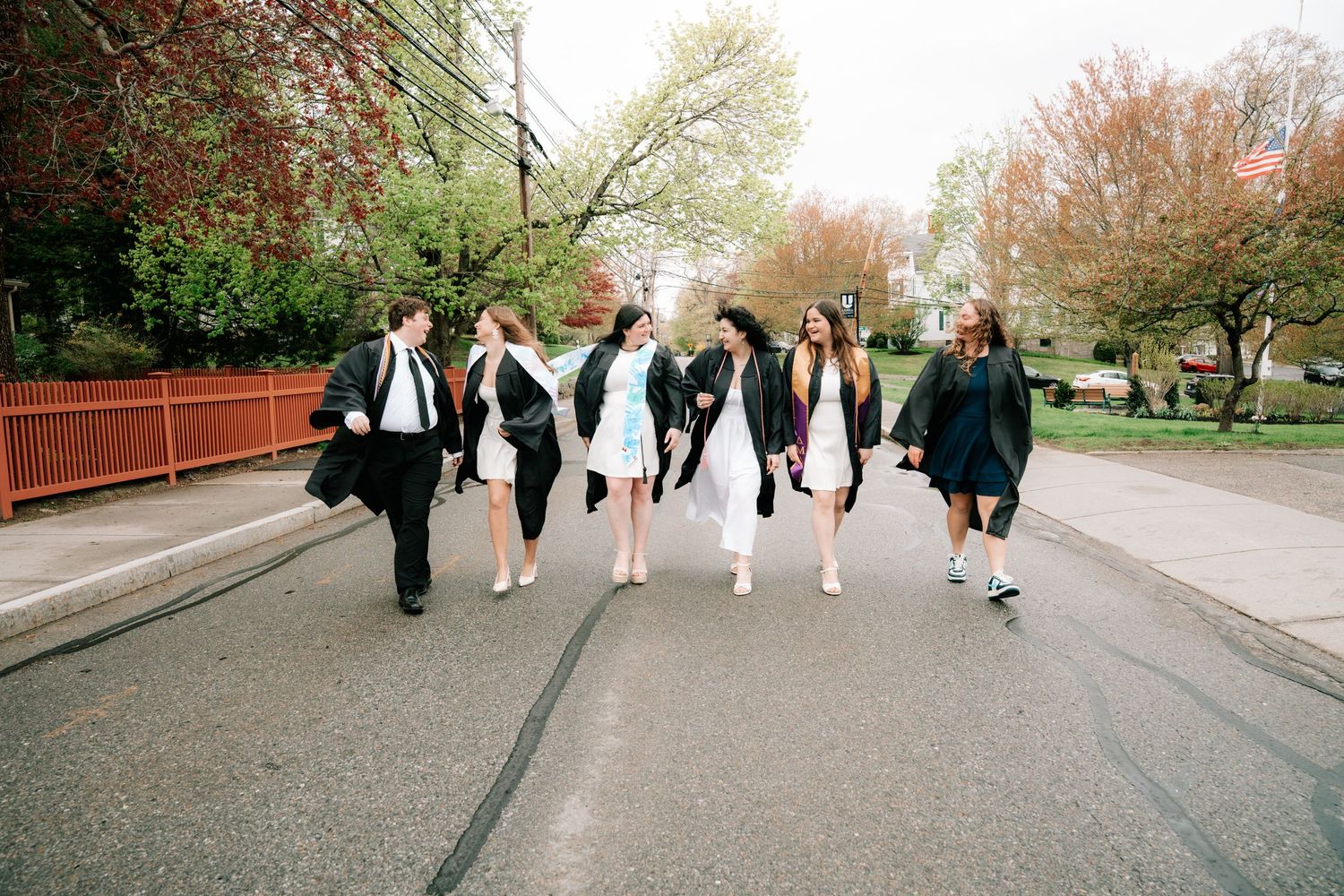 A group of graduates in black gowns walk together laughing down a tree-lined campus road.