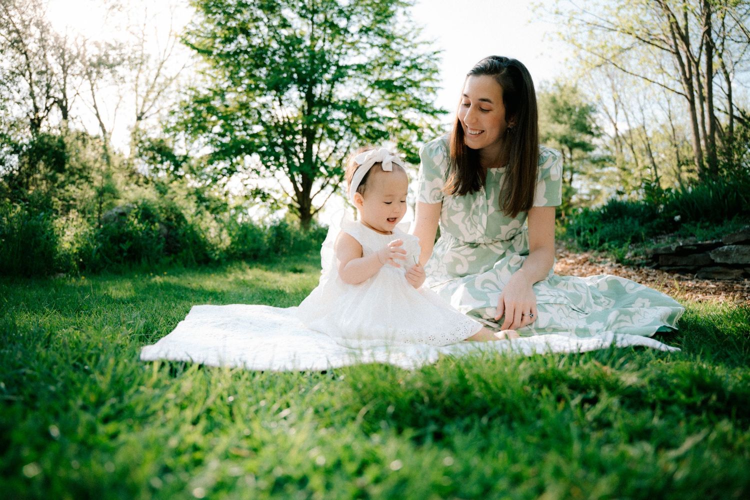 A mother and baby girl in white dress sit on a blanket in a lush green park on a sunny day.