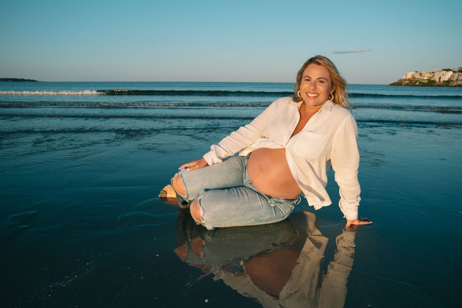Pregnant woman sitting on wet beach sand in jeans and white shirt, smiling with ocean backdrop at golden hour.