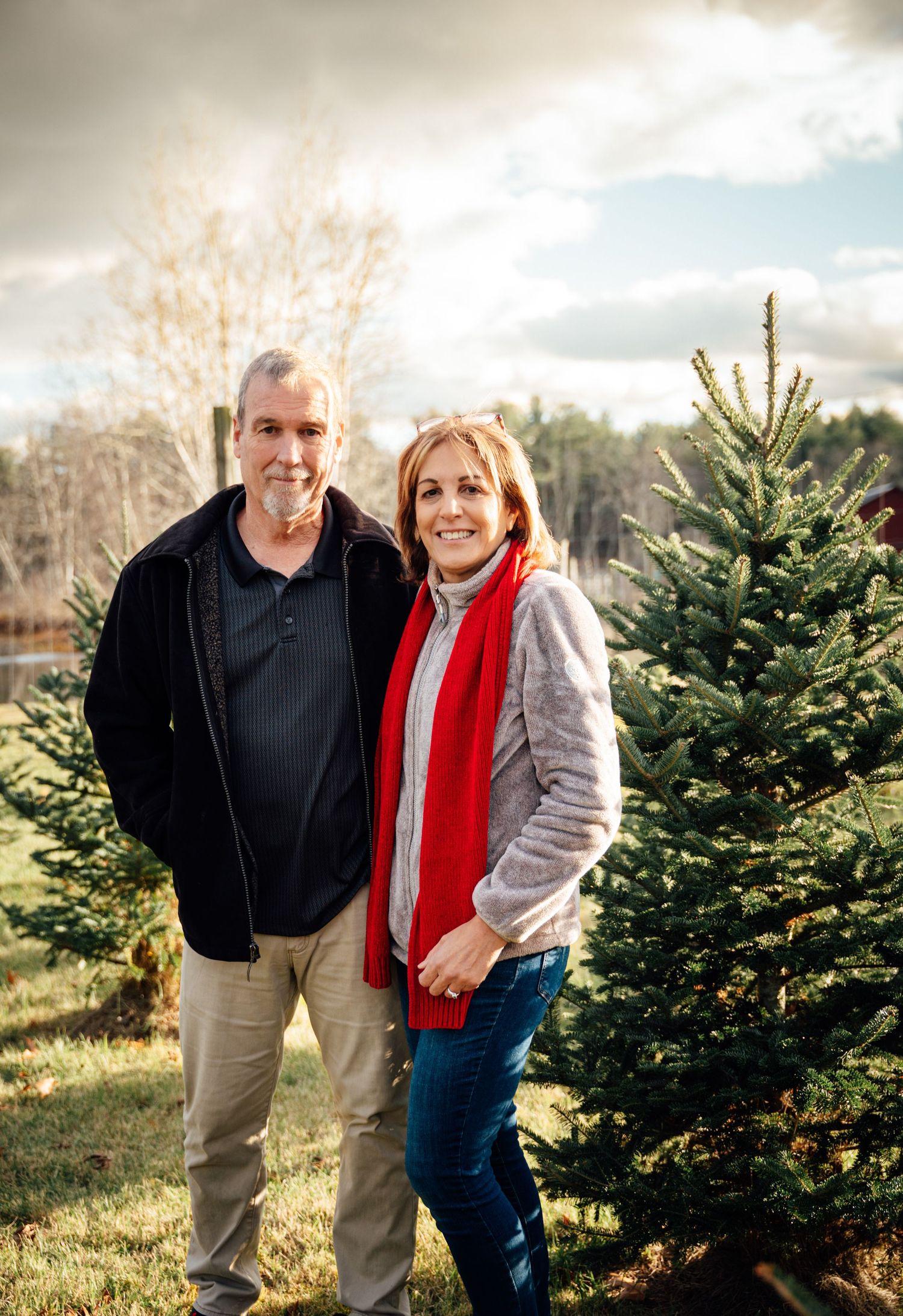 Couple smiling together beside green Christmas trees outdoors.