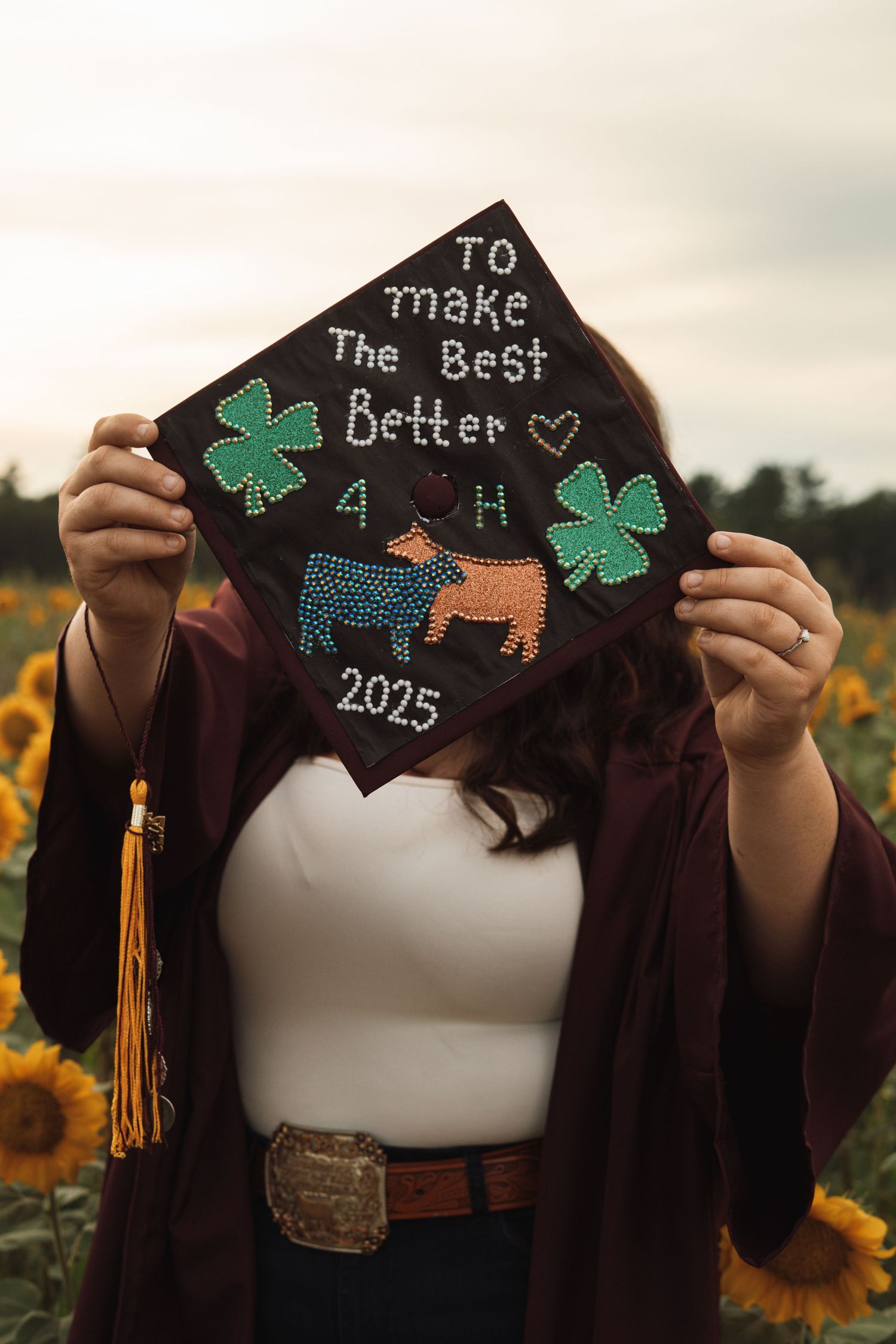 A graduate holds up a decorated black mortarboard cap reading To Make The Best Better with 4-H symbols and 2025.