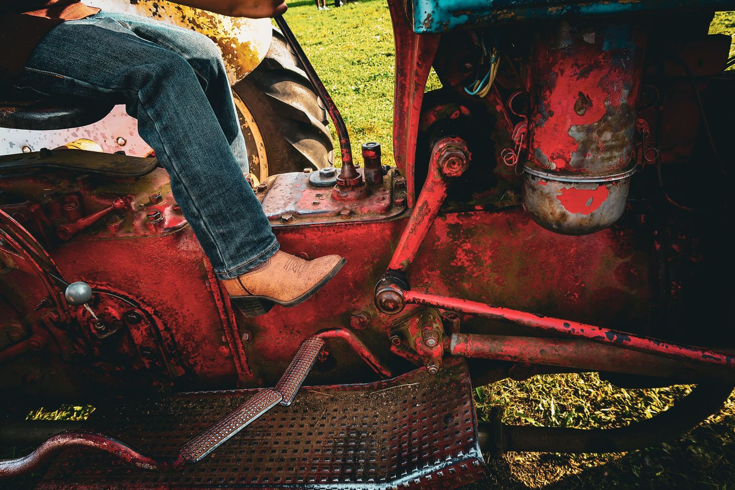 Person wearing cowboy boots sitting on a rusty vintage red tractor outdoors.