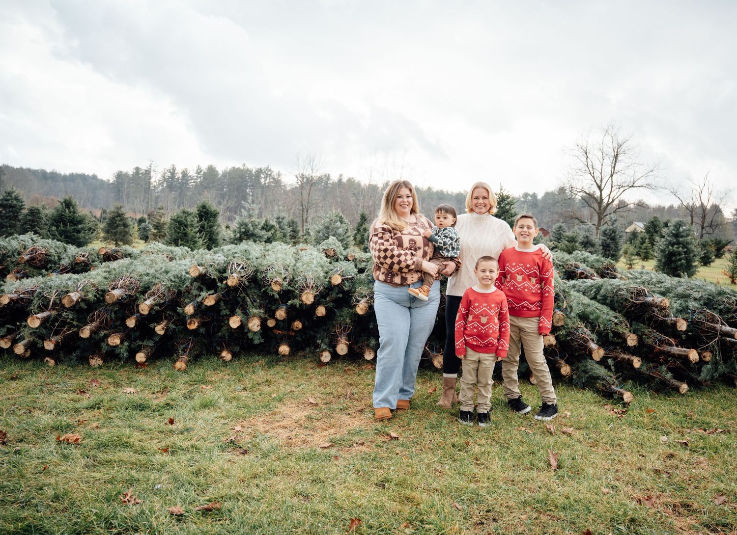 Family of five posing at a Christmas tree farm with rows of cut trees behind them.