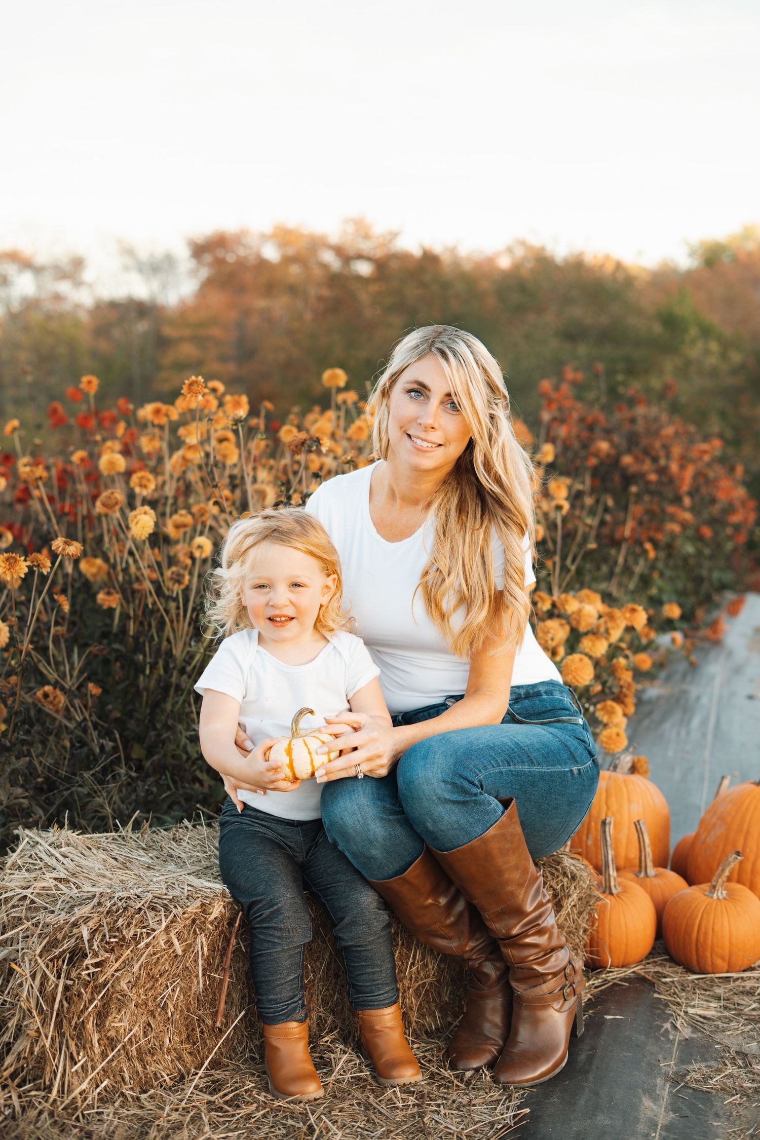 A smiling blonde mother and her toddler daughter sit on a hay bale surrounded by pumpkins and fall flowers.