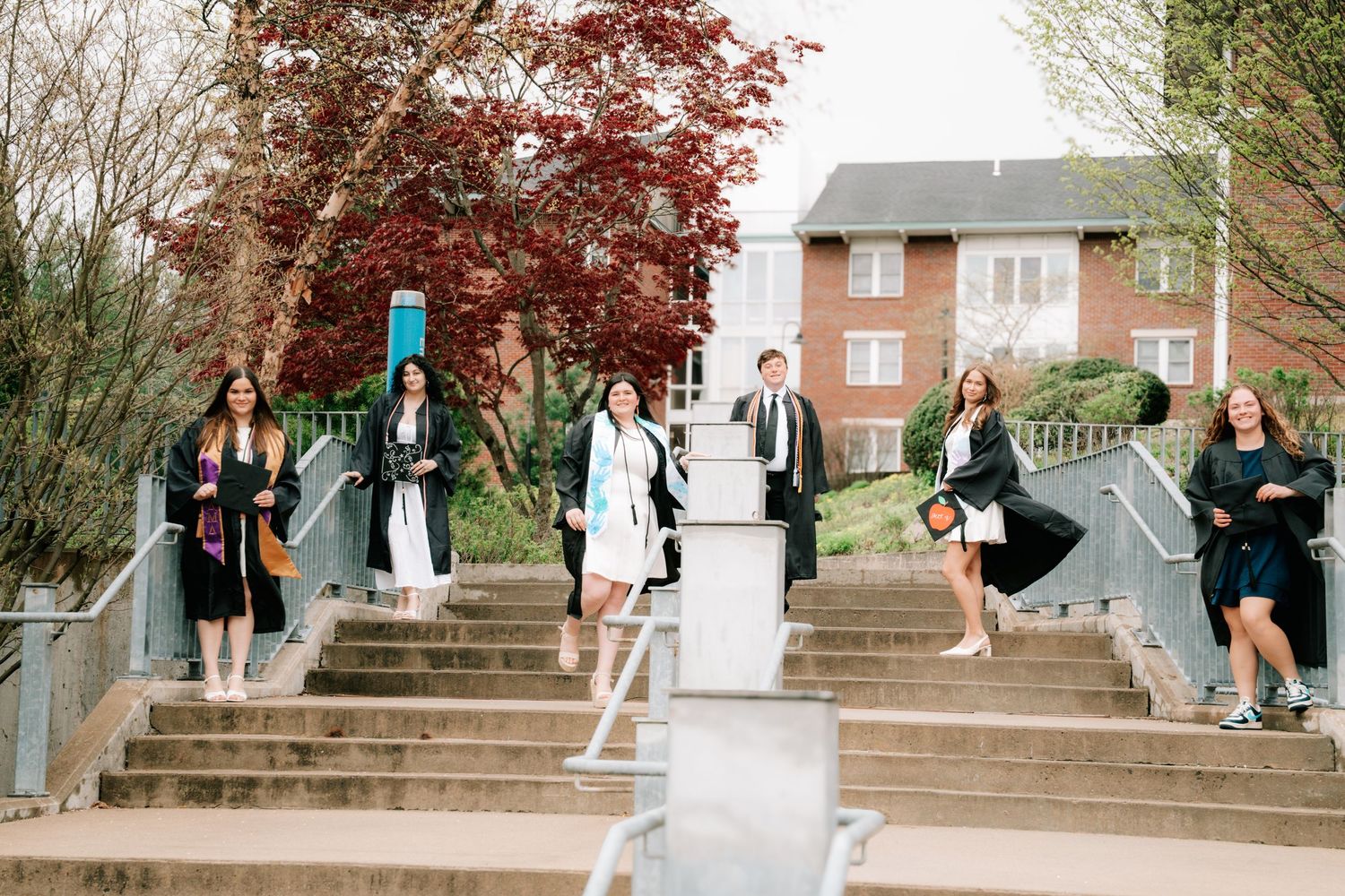 Group of graduates in black robes posing on outdoor campus staircase surrounded by spring trees.