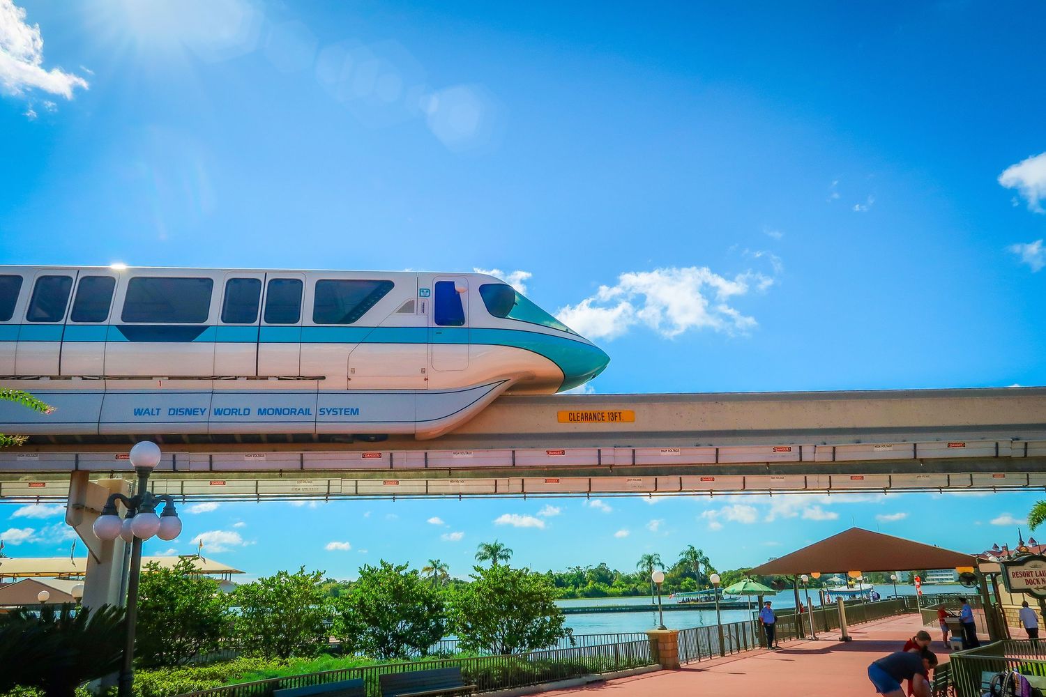 A teal and white Walt Disney World monorail glides along its elevated track under a bright blue sky.