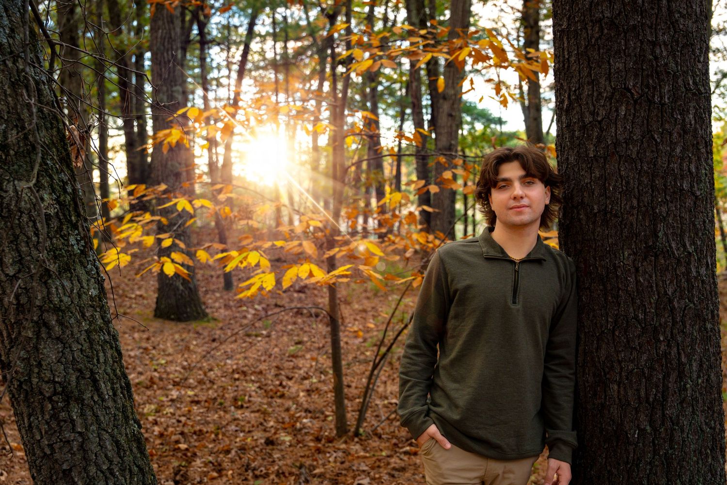 Teen boy in green pullover leans on tree in golden autumn forest at sunset.