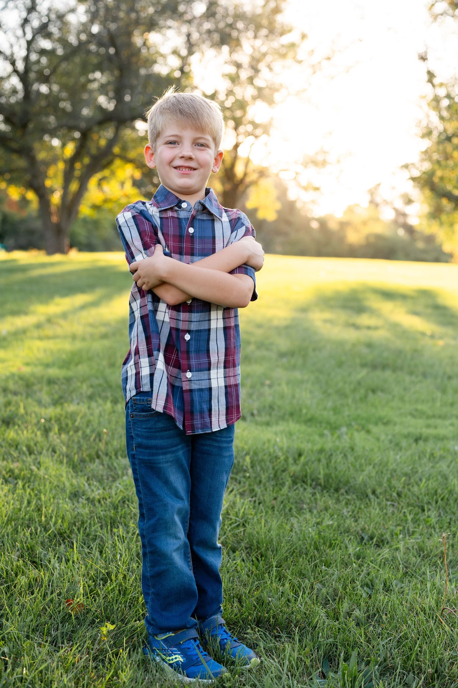 Child in plaid shirt posing with crossed arms on grassy lawn at sunset.