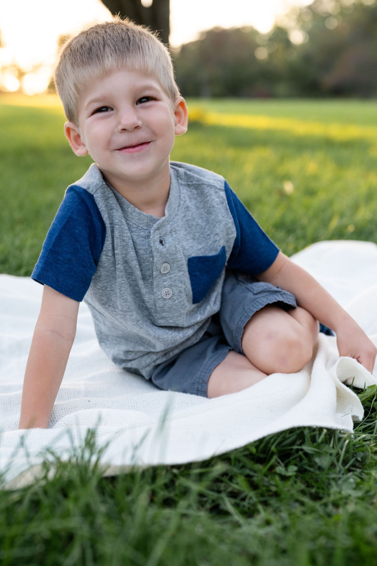 Child in grey and blue shirt sitting on white blanket in sunny field at sunset.