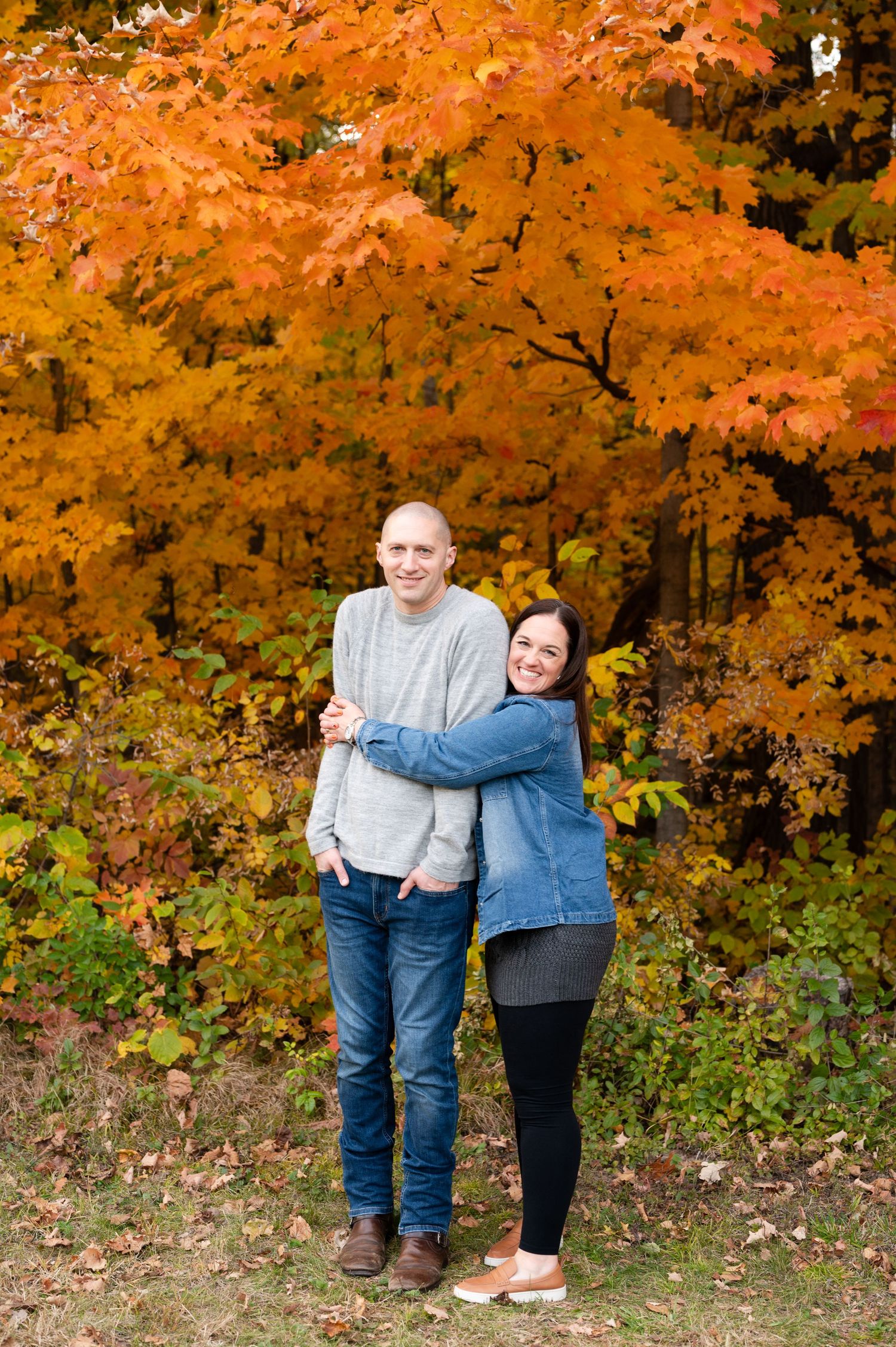Couple embracing in casual pose against backdrop of bright orange autumn leaves.