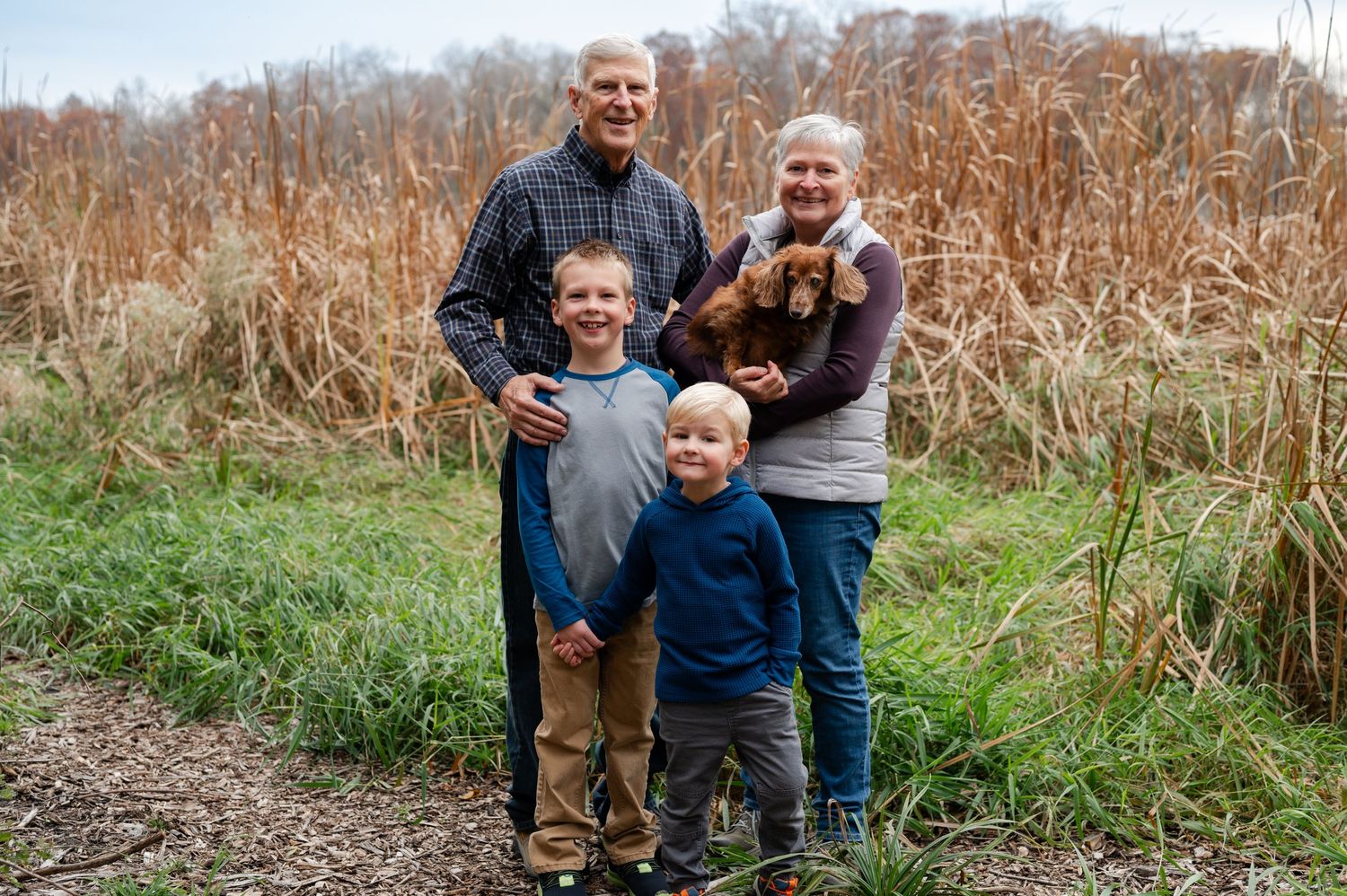 A family smiles together while standing on a nature trail with brown reeds in the background.