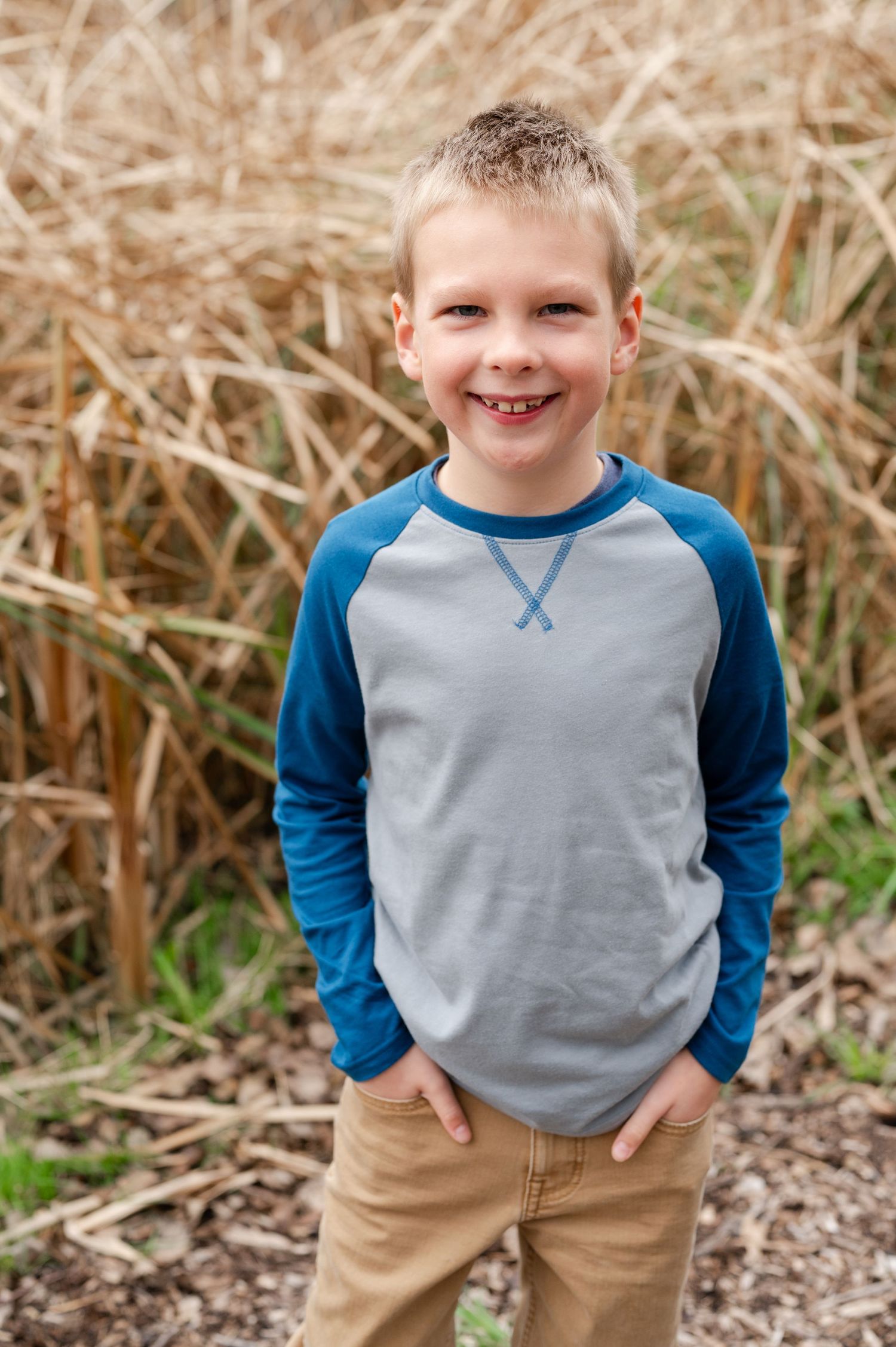Young boy in blue and gray baseball shirt smiling against natural grass backdrop.